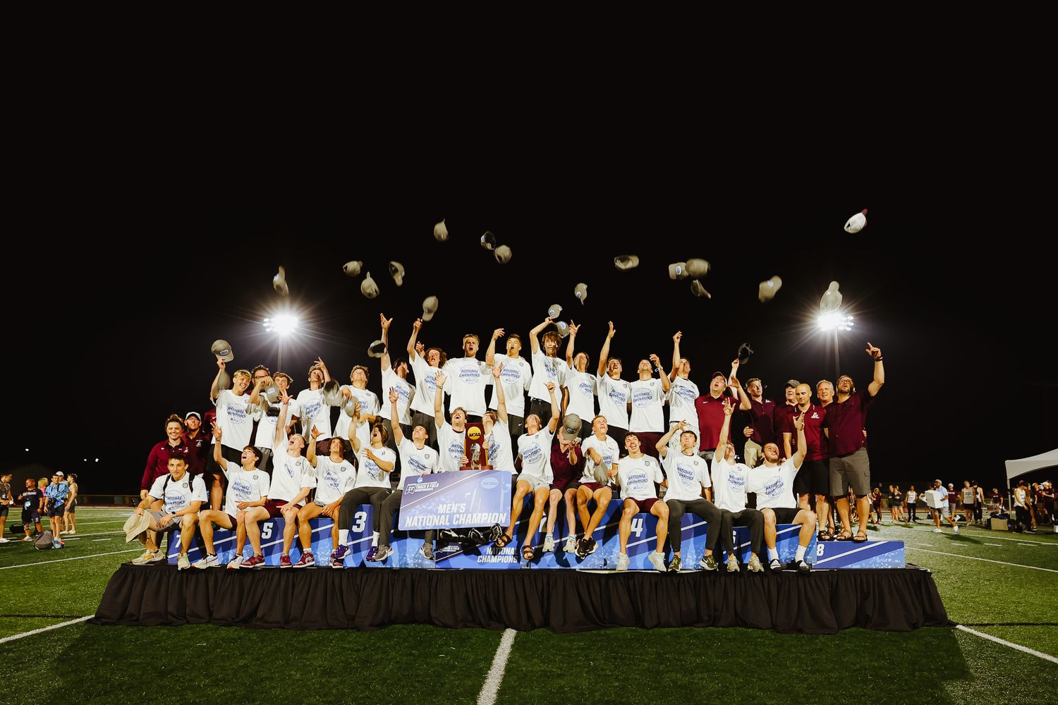Students celebrate on a soccer field at night with stadium lights illuminating their joyful group pose.