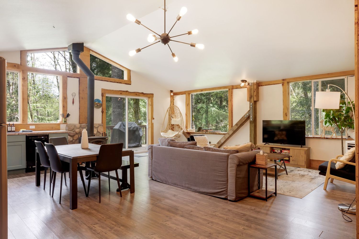 Open-concept living and dining room with vaulted ceilings, modern chandelier, large windows, hardwood floors, and natural wood accents.