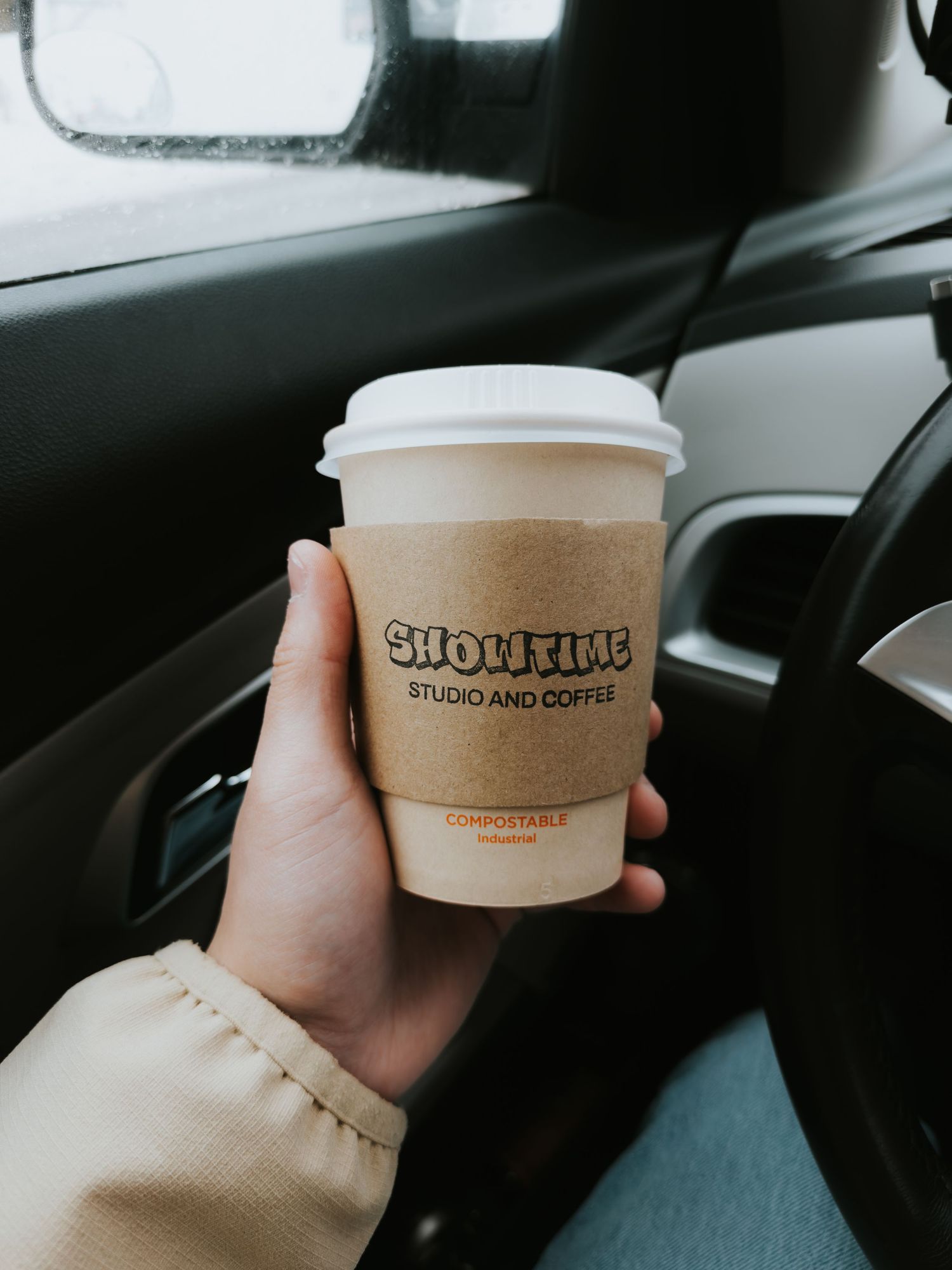 A hand holds a disposable coffee cup with a brown sleeve while sitting in a car.