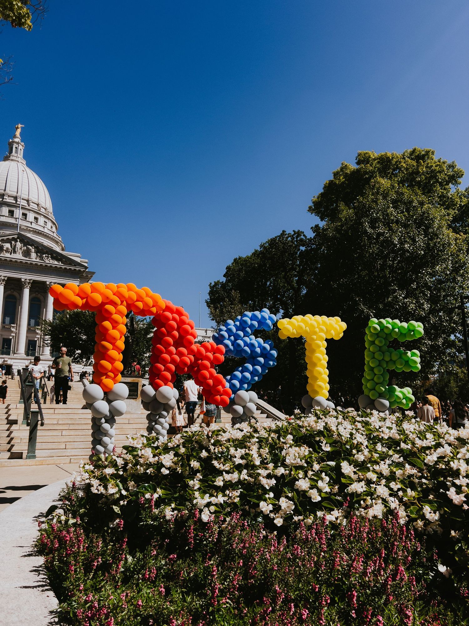 Colorful balloon arch installation decorates an outdoor garden path with white flowers and architectural elements in background.