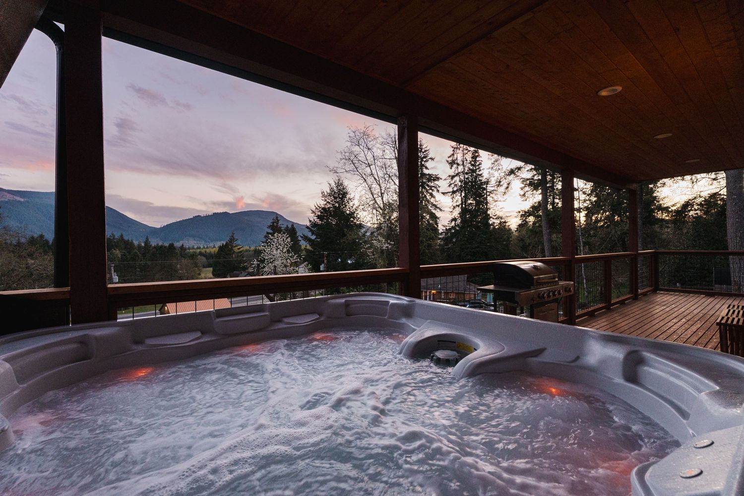 Bubbling hot tub on a wooden deck with stunning mountain and forest views at dusk.