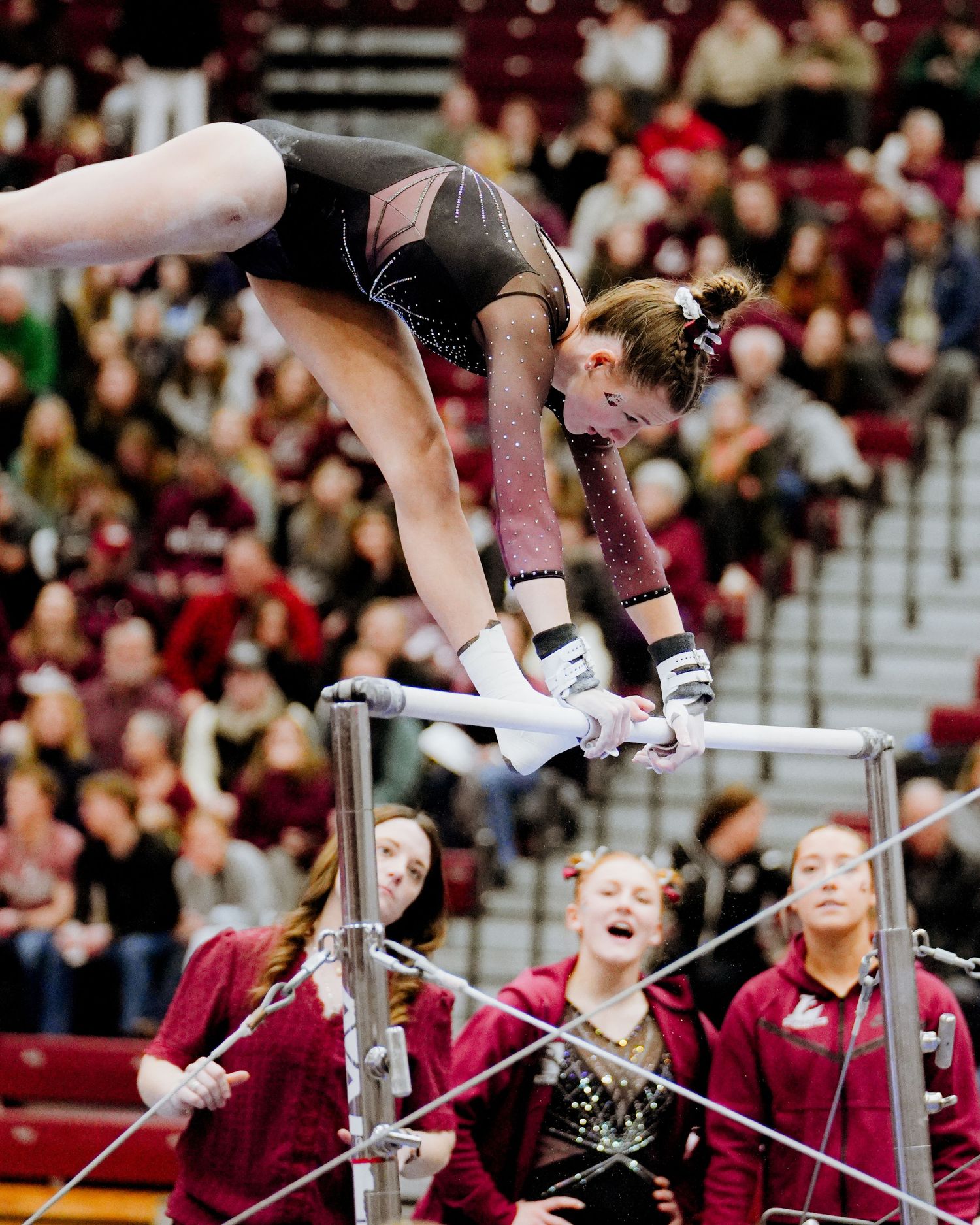 Gymnast performs a high-flying uneven bars routine during a collegiate competition with maroon-clad spectators watching.