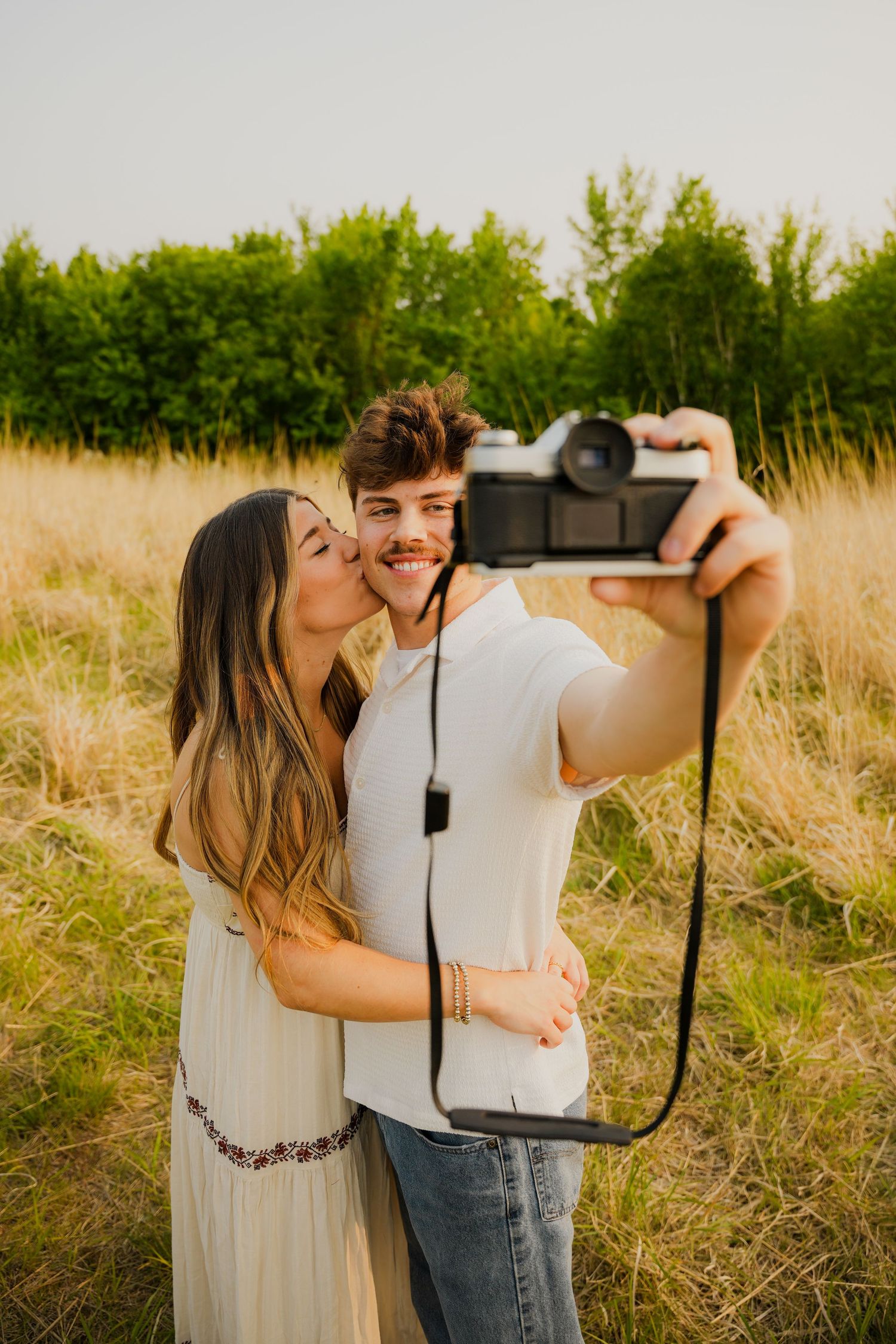 A couple takes selfies together in a golden wheat field during sunset while sharing affectionate moments.