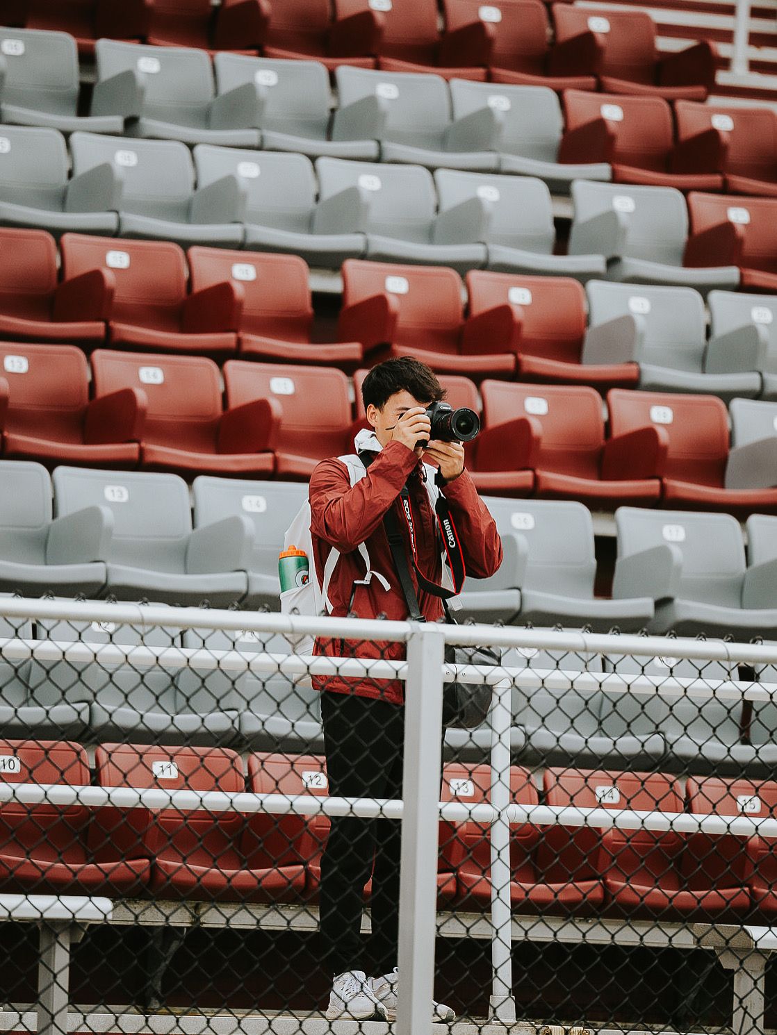 Empty stadium seats in red and white with chain link fence visible in foreground.