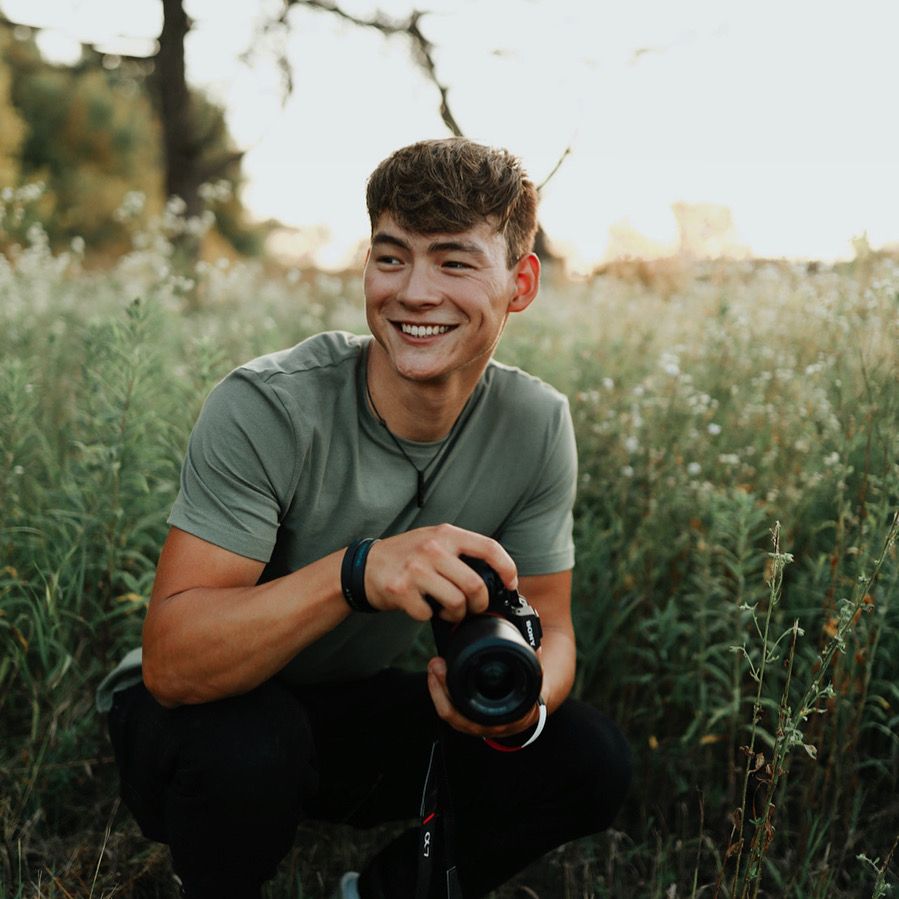Photographer holding camera smiles while crouching in wildflower field at golden hour.
