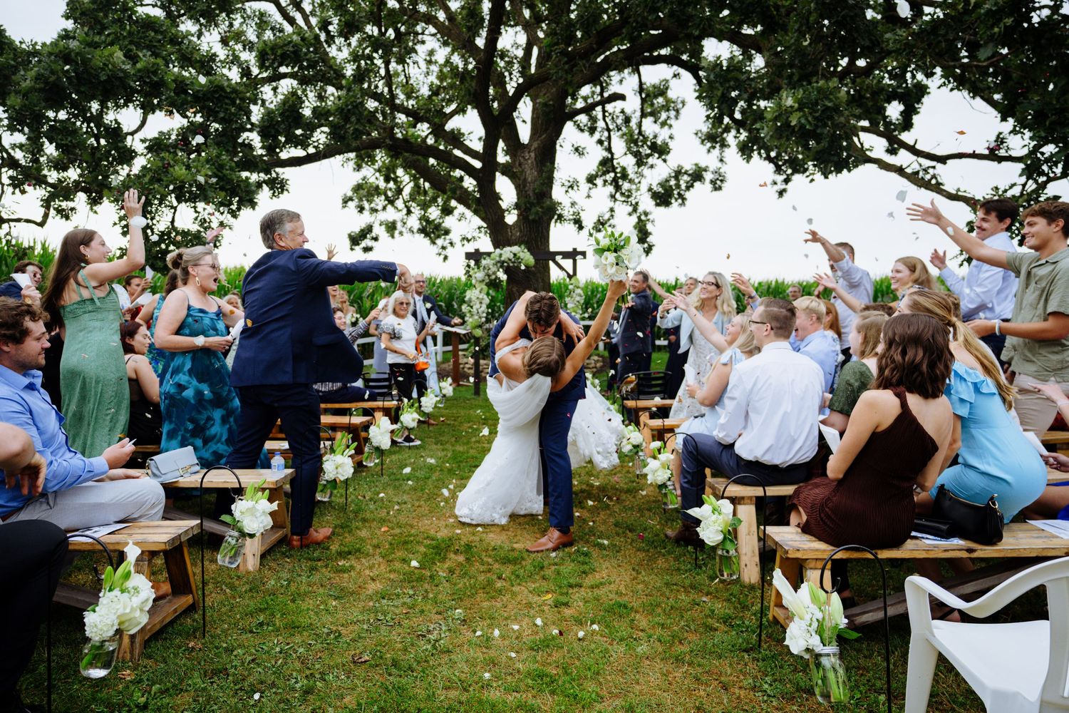 Wedding guests sit outdoors in white chairs during an intimate garden ceremony surrounded by lush greenery.