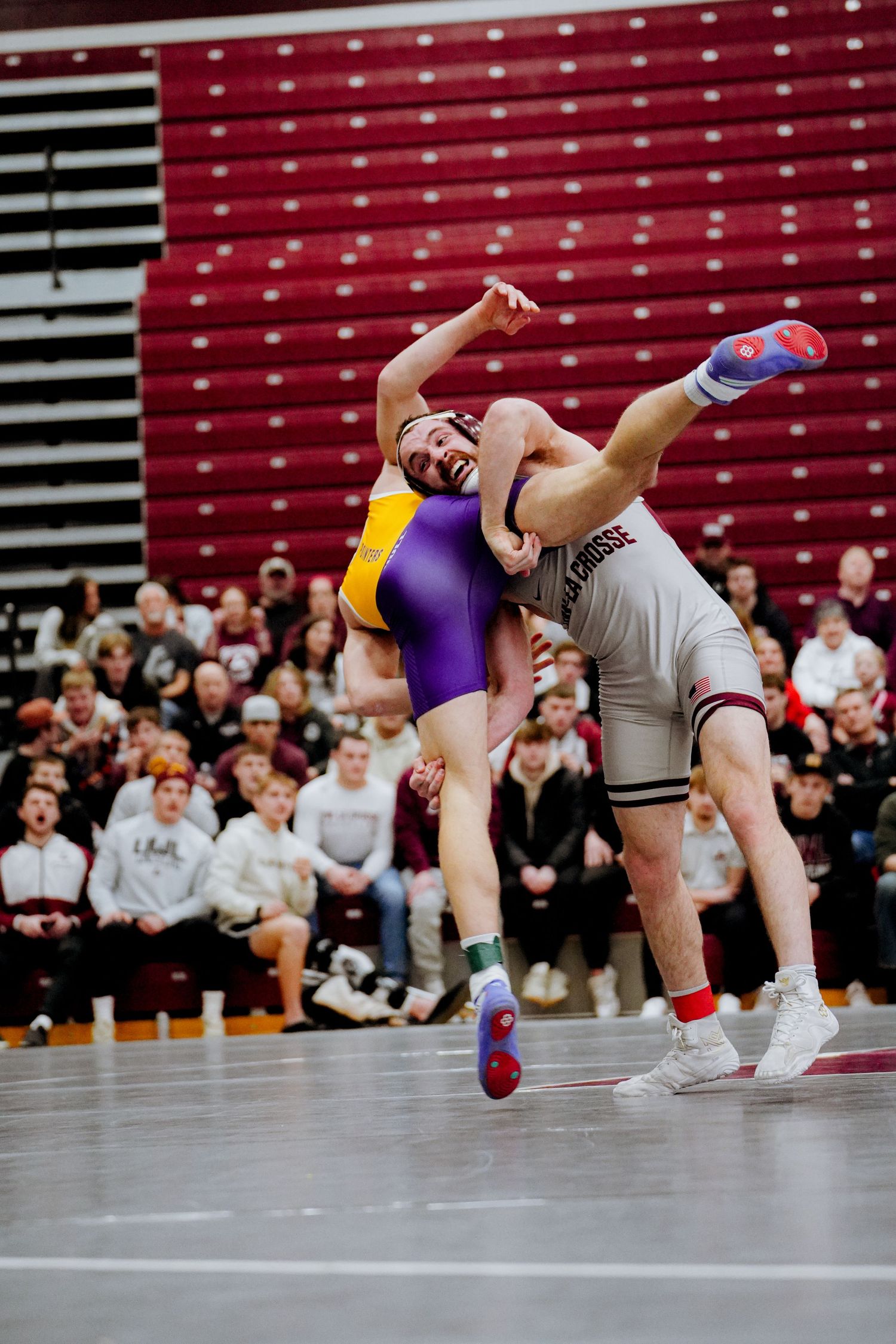 Two wrestlers compete in an intense match on the mat during a college wrestling competition with spectators watching.