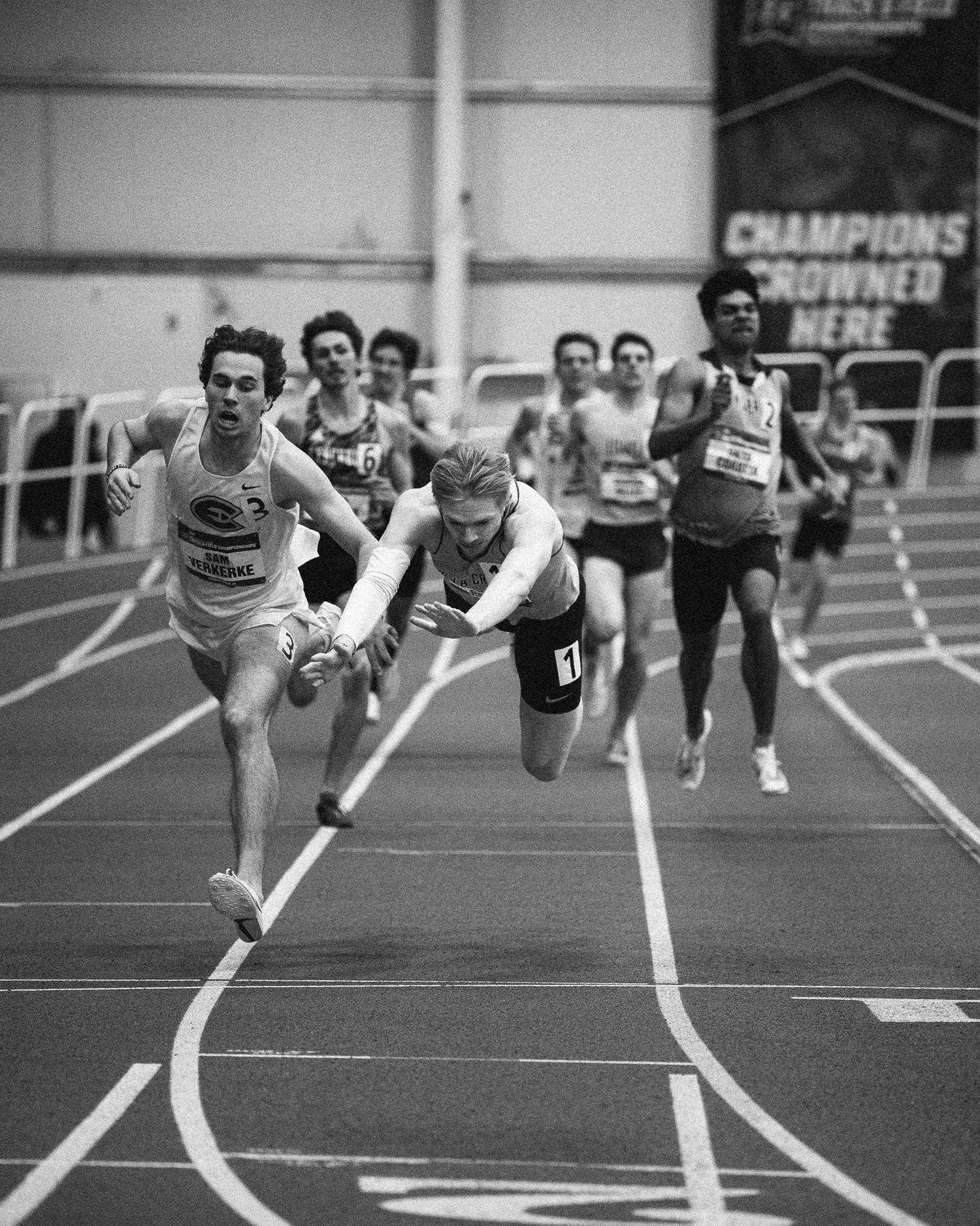 Black and white photo of runners diving across finish line during indoor track meet.