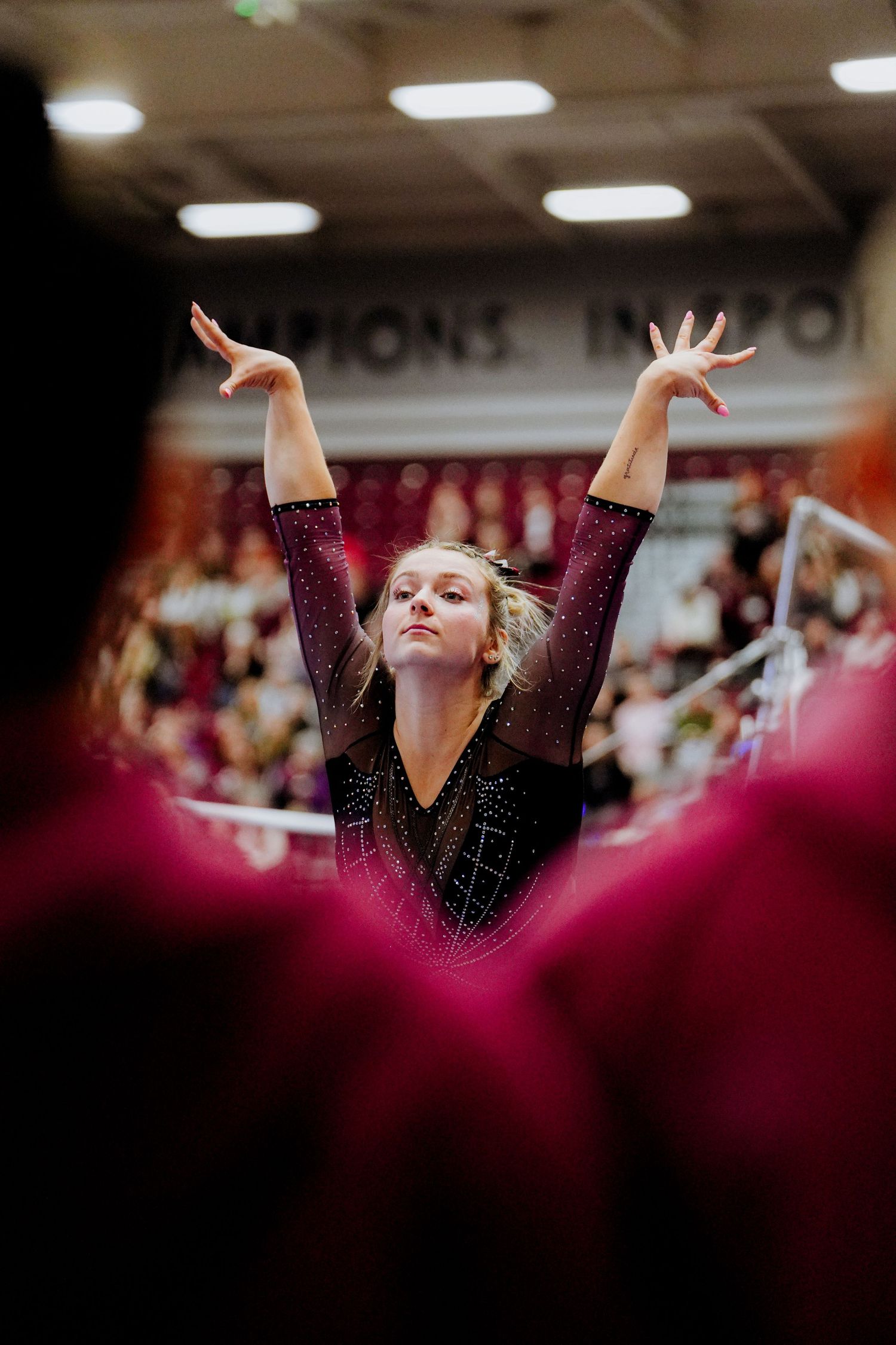 Gymnast in black sparkly leotard performs graceful arm movements during floor routine with crowd in background.
