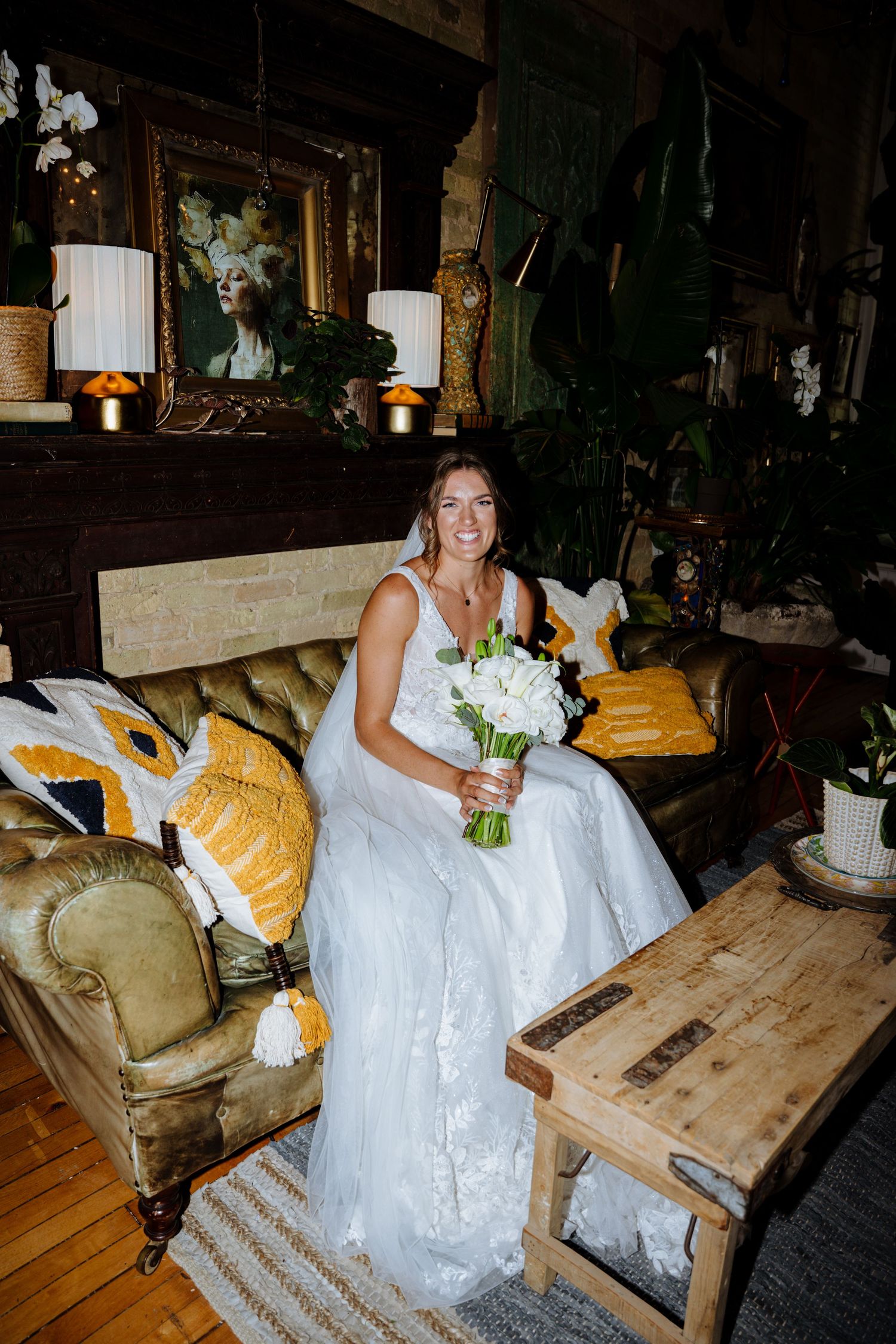 A person in a white wedding dress sits on a rustic wooden bench with yellow cushions in a dimly lit room.