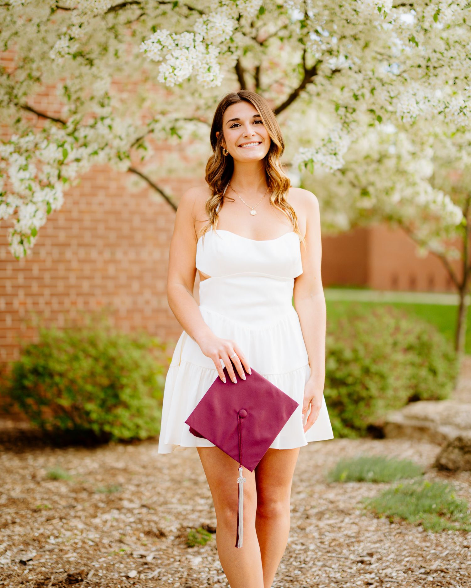 Graduate in white dress holding burgundy cap under blooming spring trees.