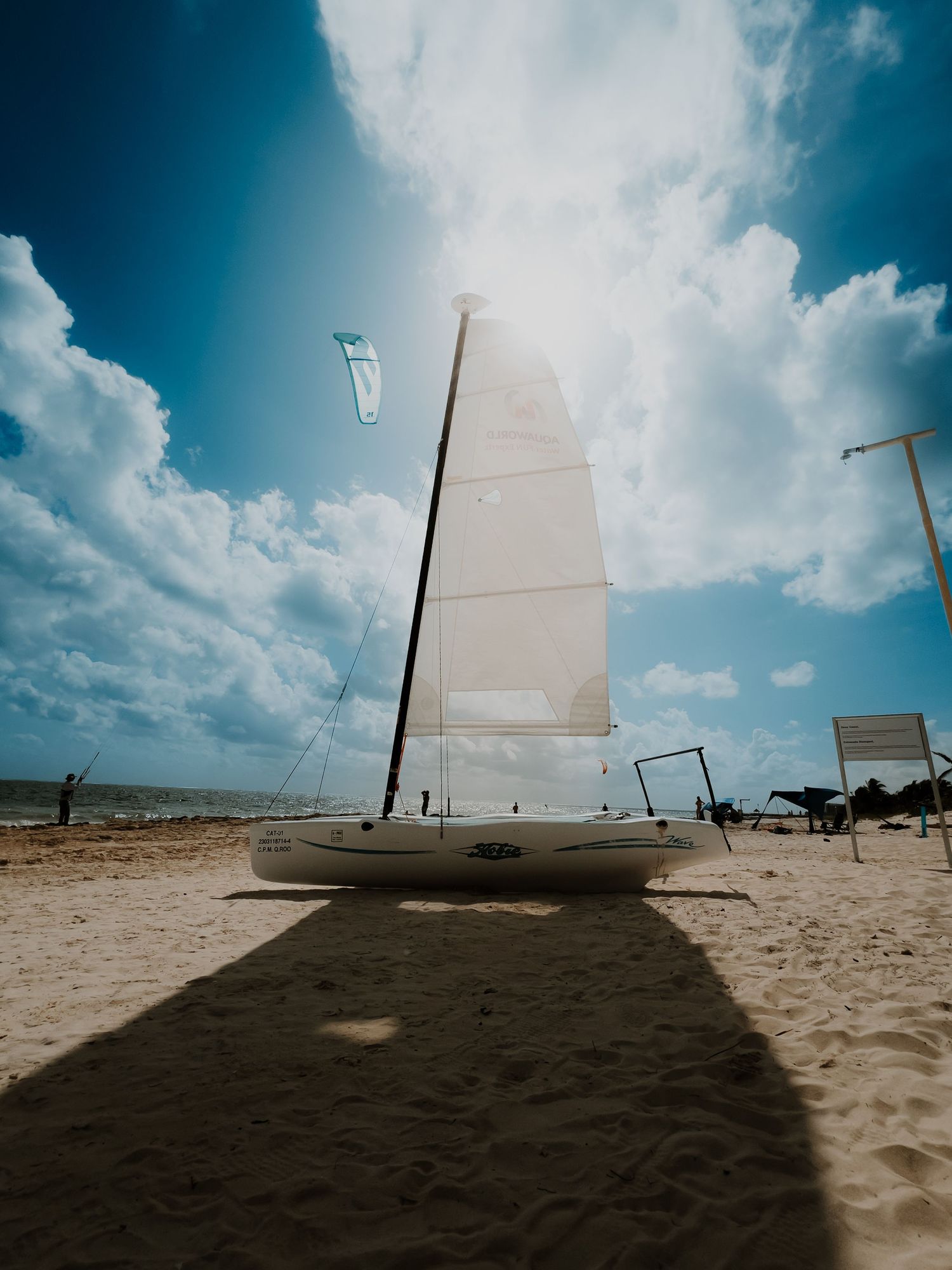 A white sailboat casts a long shadow on a sandy beach against a bright blue sky with puffy white clouds.