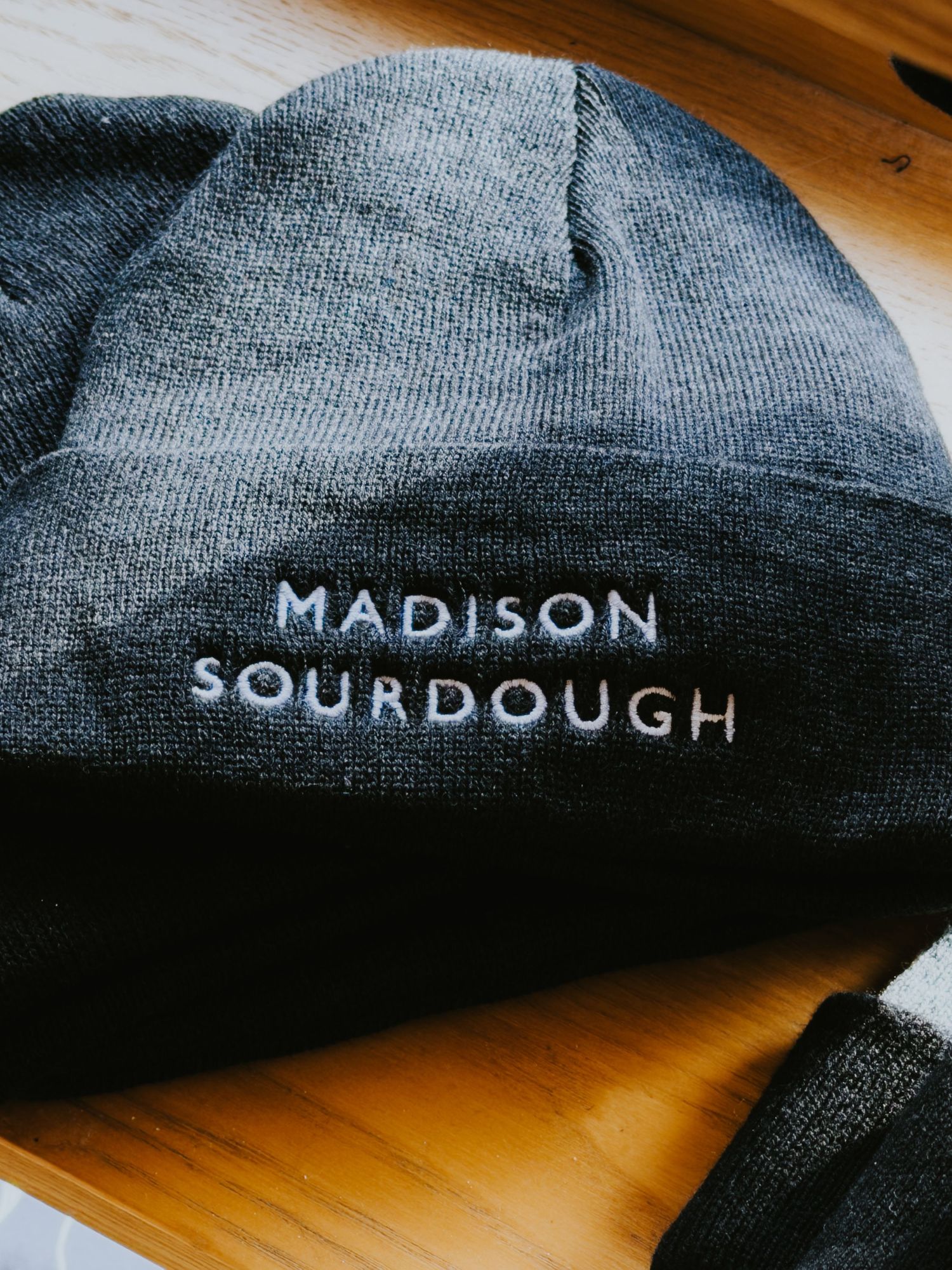 A black knit beanie with 'Madison Sourdough' text embroidered in white on a wooden surface.