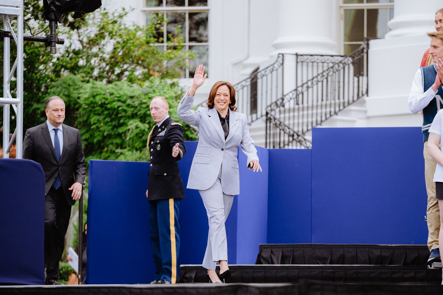 A person in a light gray suit walking on a stage with blue panels and white columns.