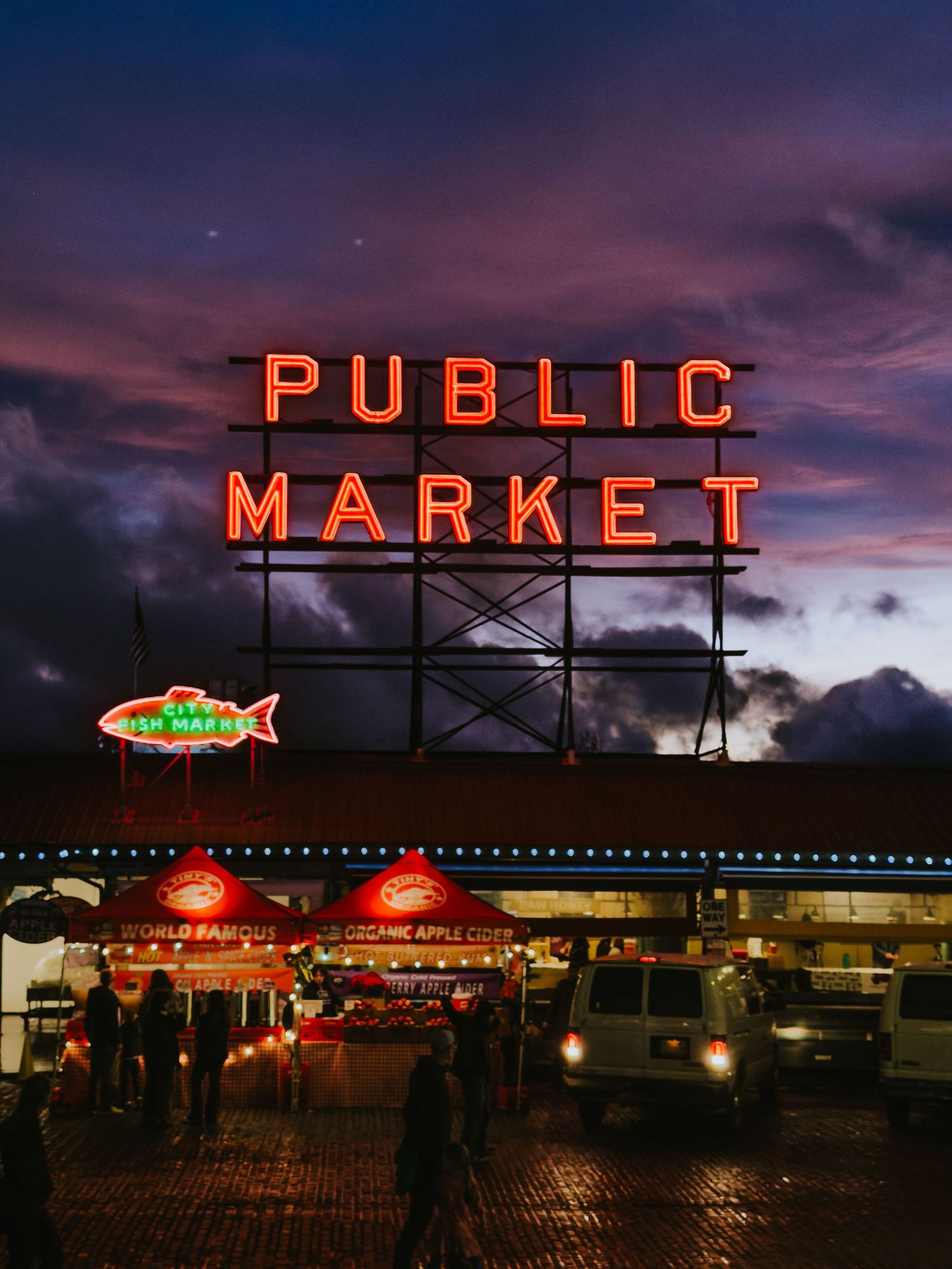 Illuminated red neon 'PUBLIC MARKET' sign glows against a dramatic purple sunset sky over Pike Place Market in Seattle.