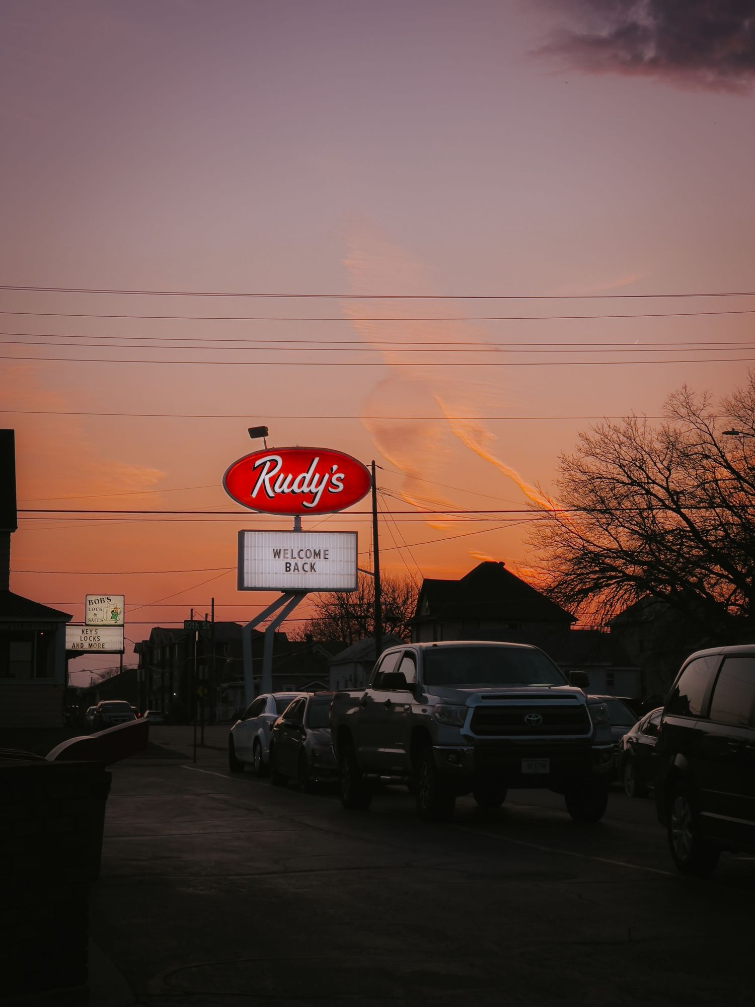 Illuminated Rudy's restaurant sign glows against a pink sunset sky with parked cars silhouetted below.