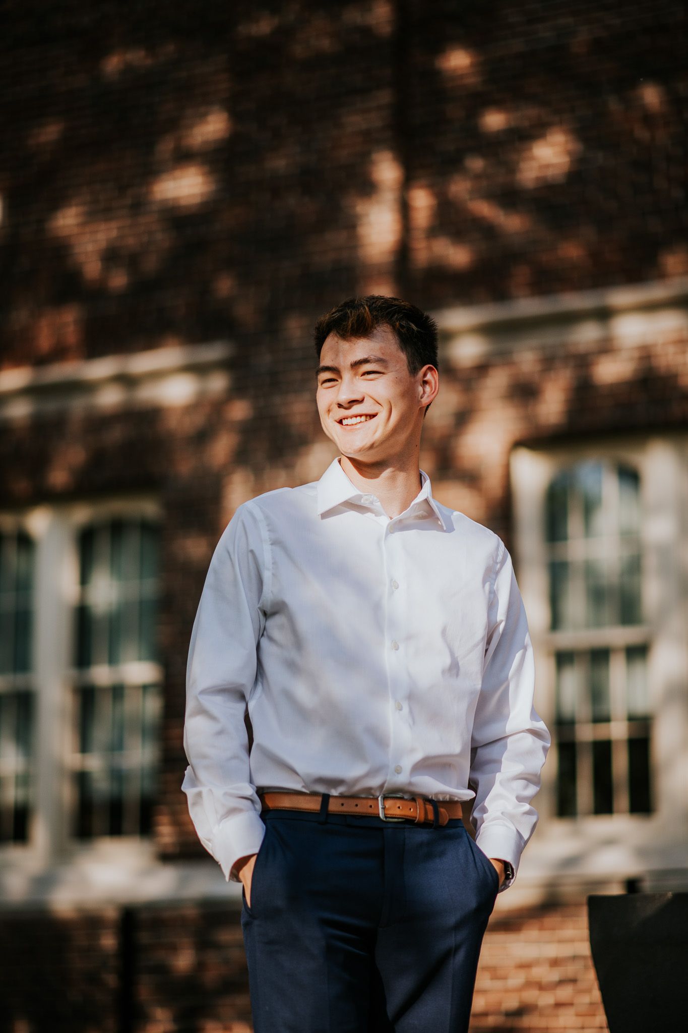 A person wearing a crisp white button-down shirt and navy pants stands in dappled sunlight with a warm smile.