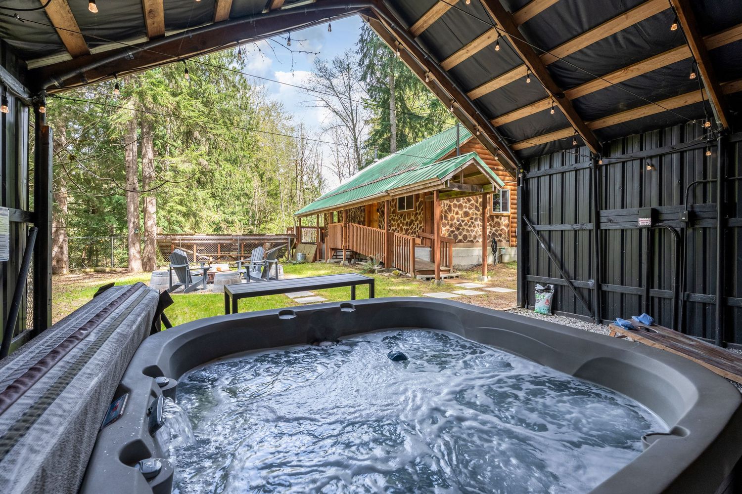 Bubbling hot tub under covered patio with rustic log cabin and lush forest backdrop.