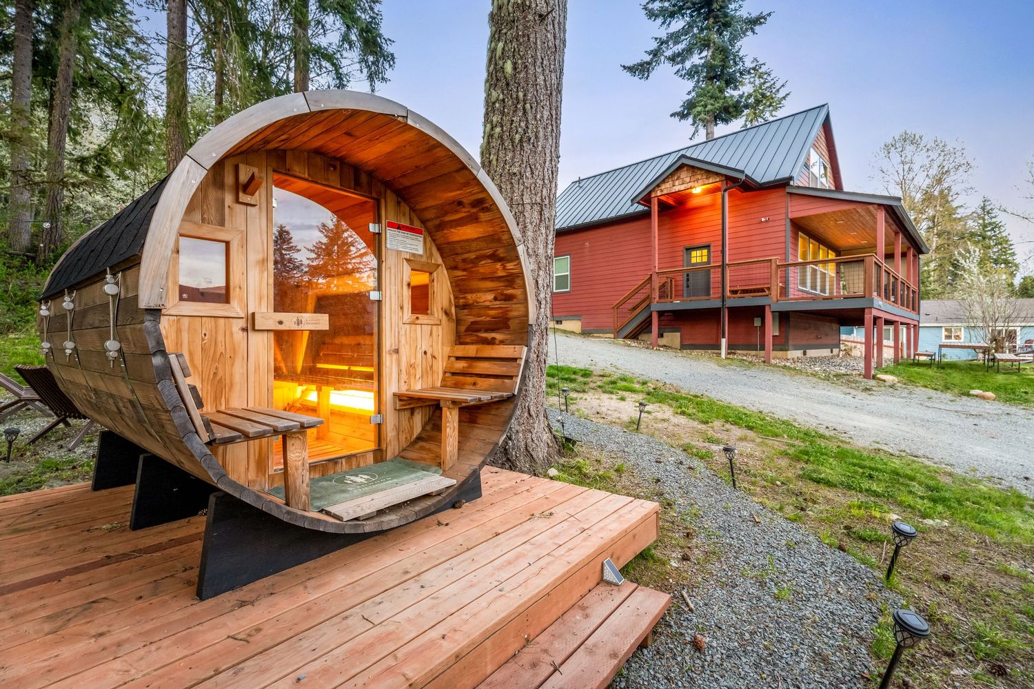 Barrel sauna on wooden deck beside tall tree with red cabin lodge in background surrounded by evergreen forest.