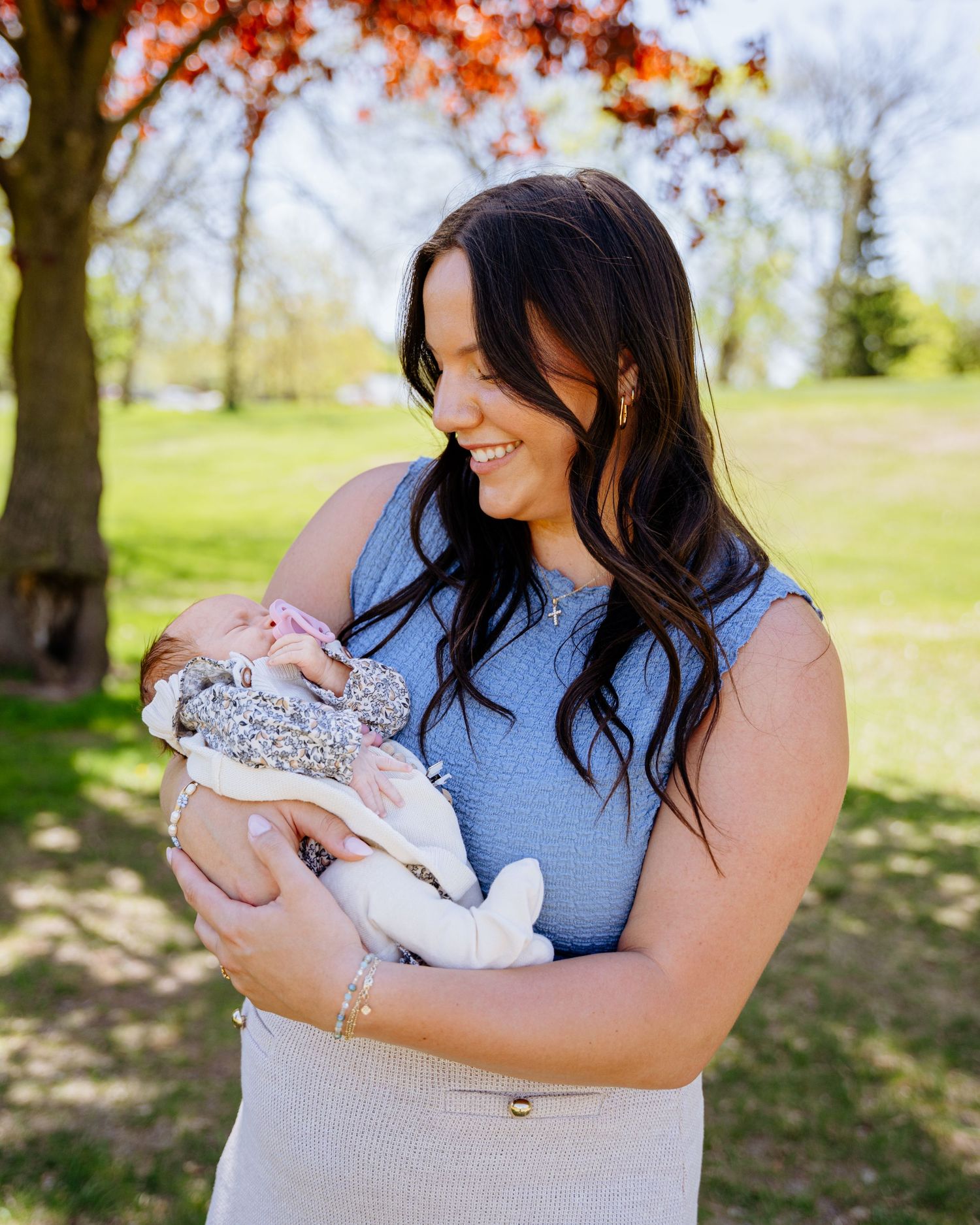 A woman in a blue dress holds a swaddled newborn baby outdoors on a sunny spring day with autumn leaves in background.