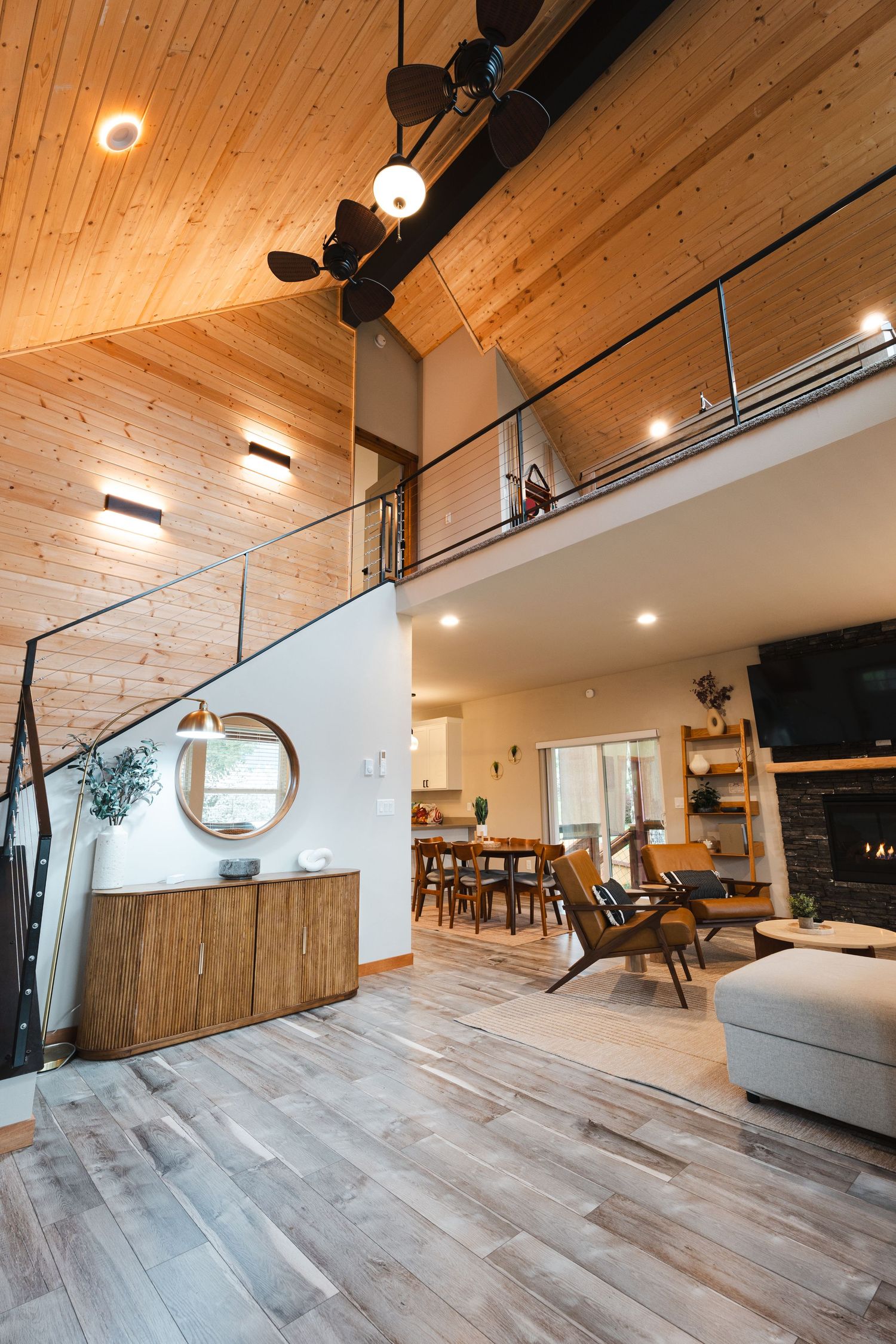 Modern open-concept living room with wood ceiling, loft, staircase, hardwood floors, and stone fireplace.