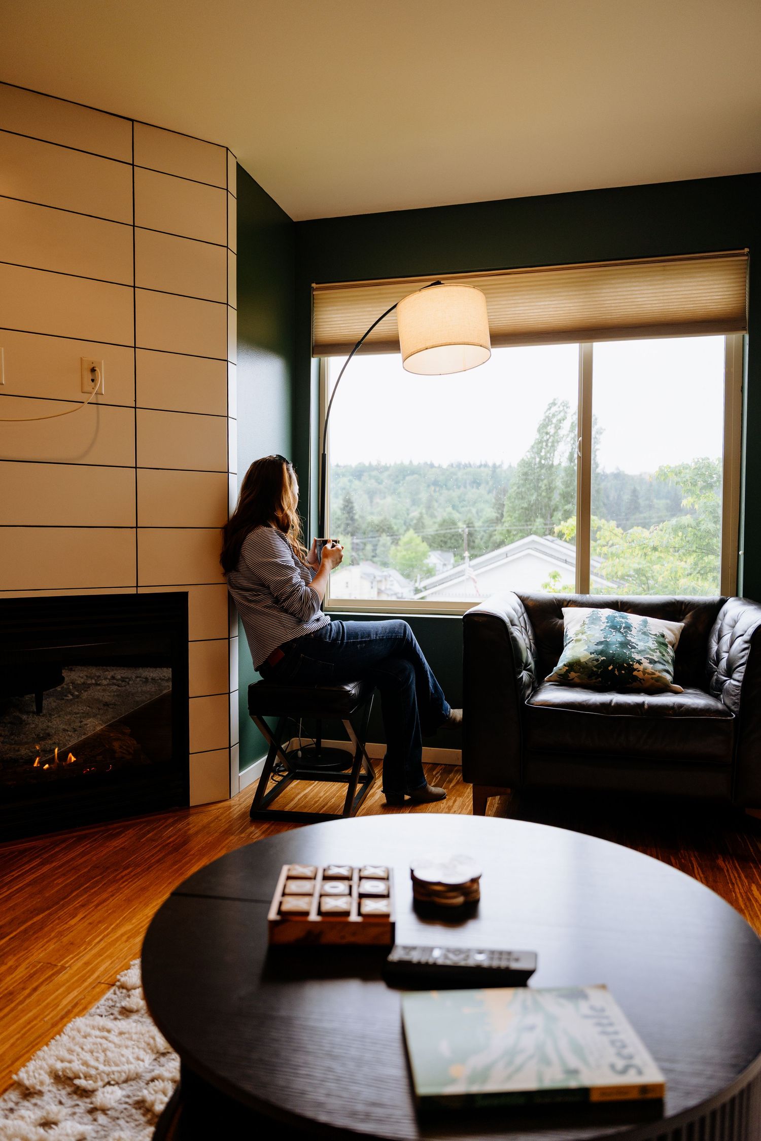 Woman sitting by window in modern living room with fireplace, floor lamp, and scenic view, holding a mug.