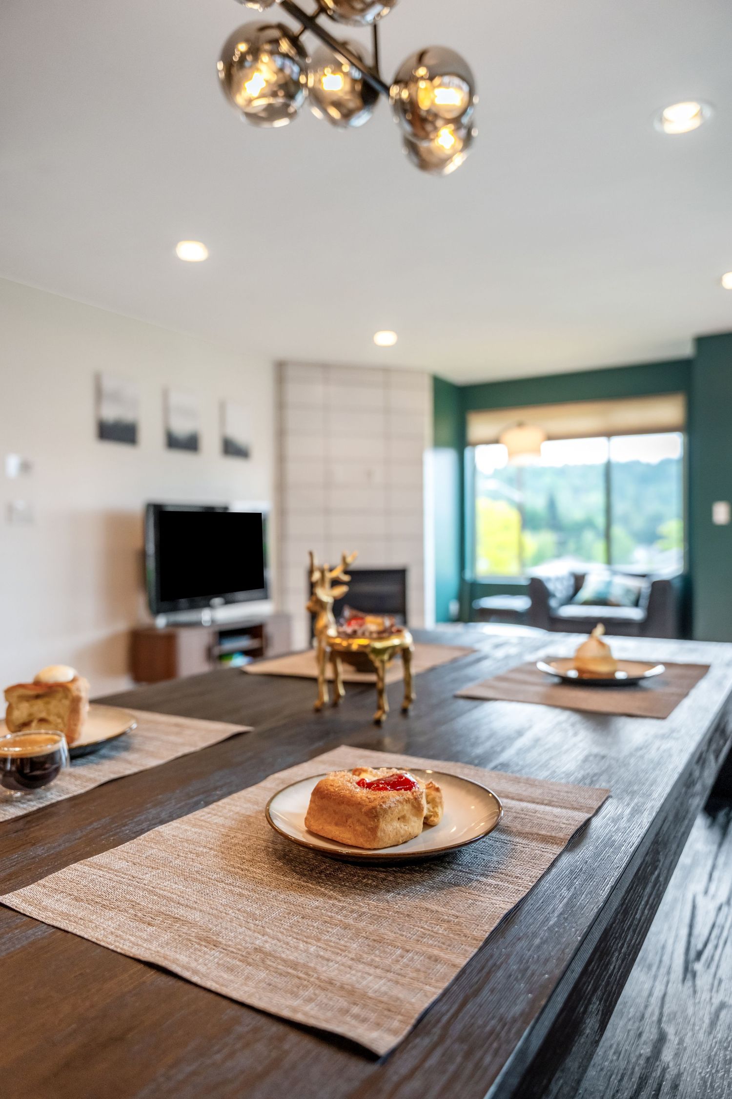 Modern open-plan dining area with dark wood table, pastry on plate, gold deer decor, TV, and green accent wall in background.