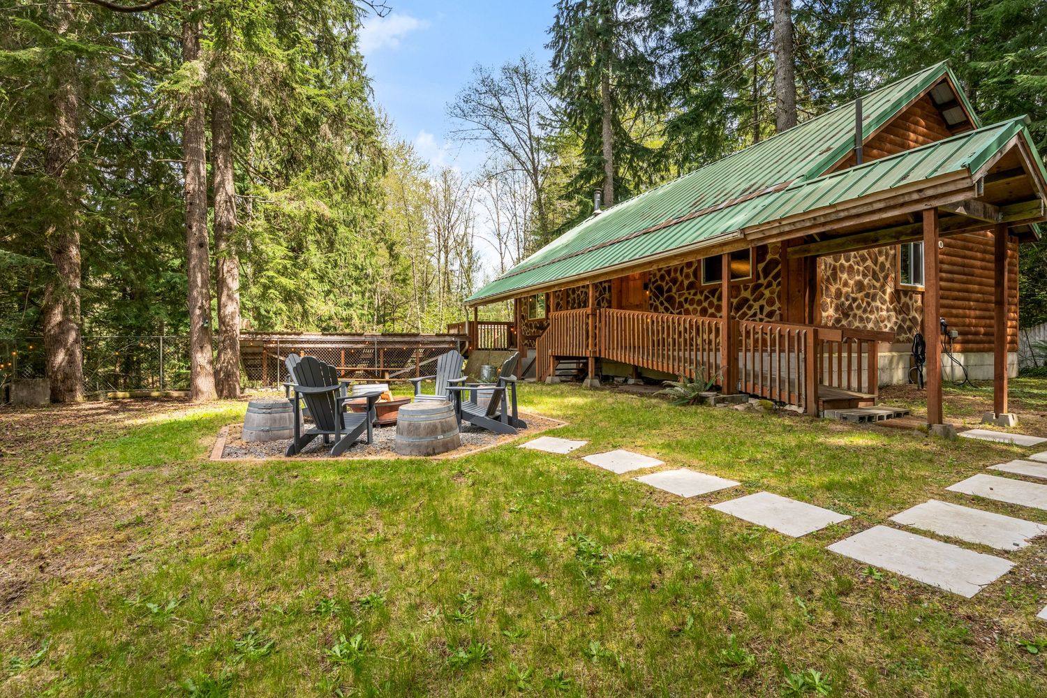 Rustic log cabin with green metal roof, fire pit seating area, and stepping stone path surrounded by tall evergreen trees.