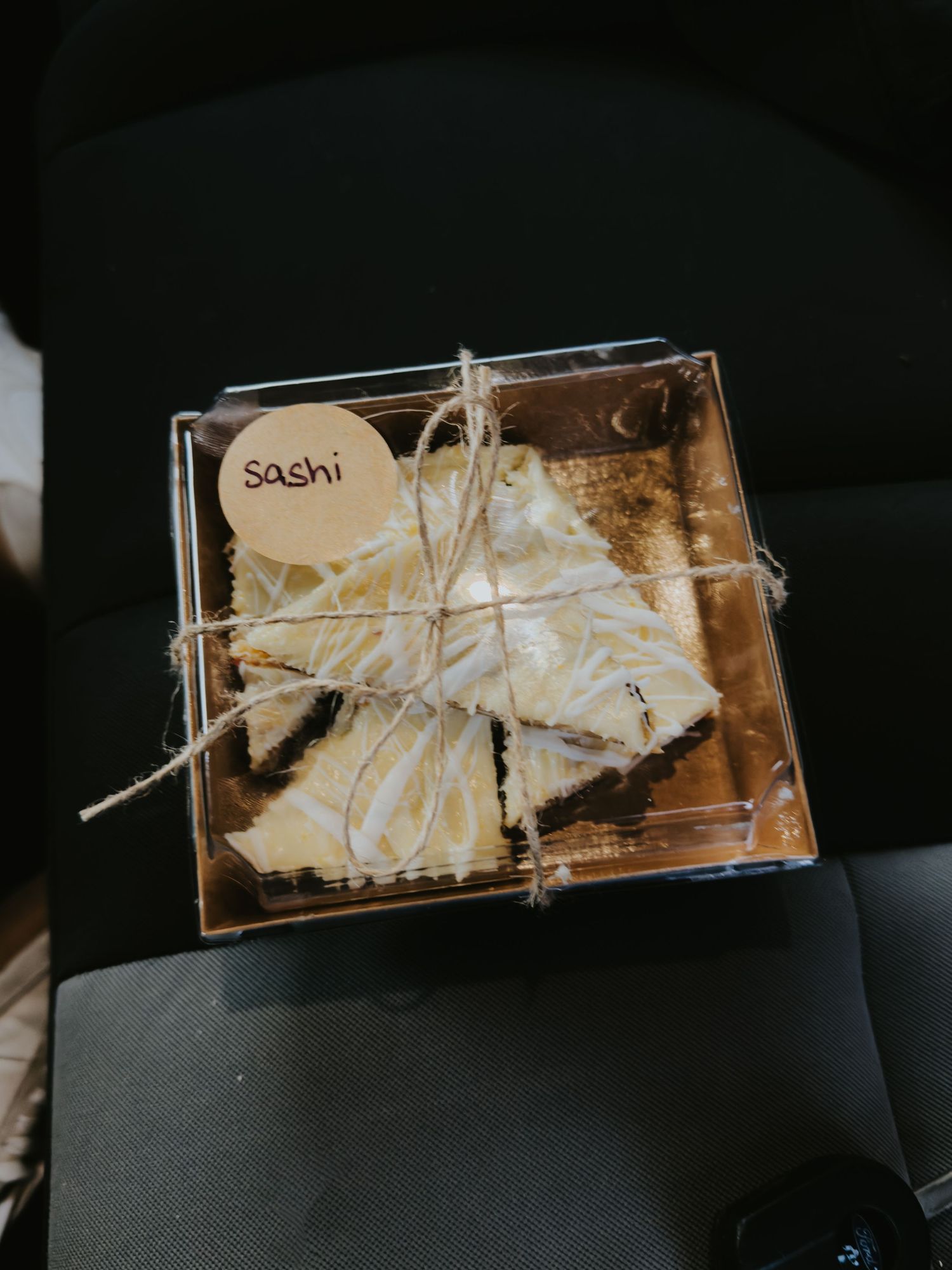 A box of sushi wrapped with twine and labeled with a simple sticker sits on a dark surface.