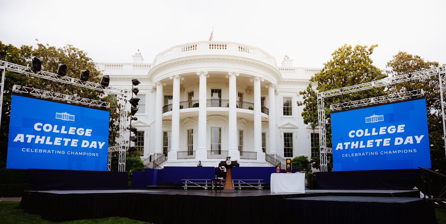 White House exterior with two large blue screens displaying 'College Athlete Day' on the South Lawn.