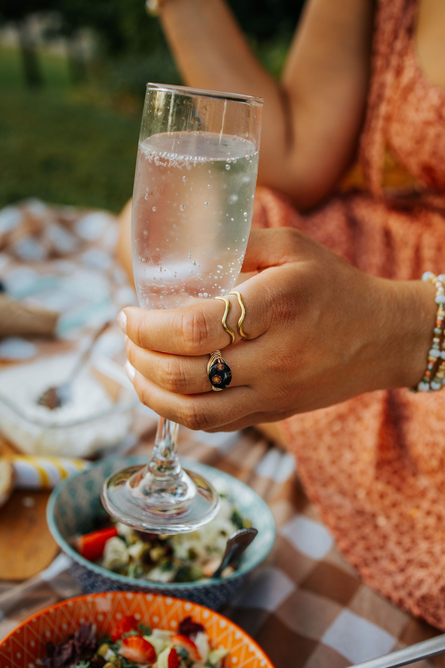 Hand holding a glass of sparkling beverage at an outdoor dining table with colorful dishes and fresh strawberries.