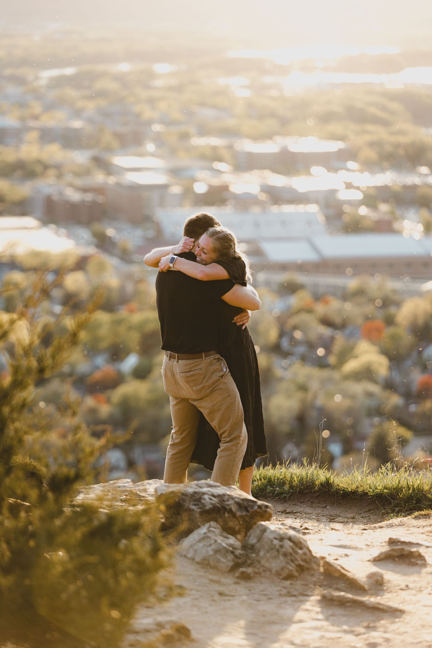 Two figures embrace while sitting on a rocky overlook at sunset with a scenic town vista in the background.