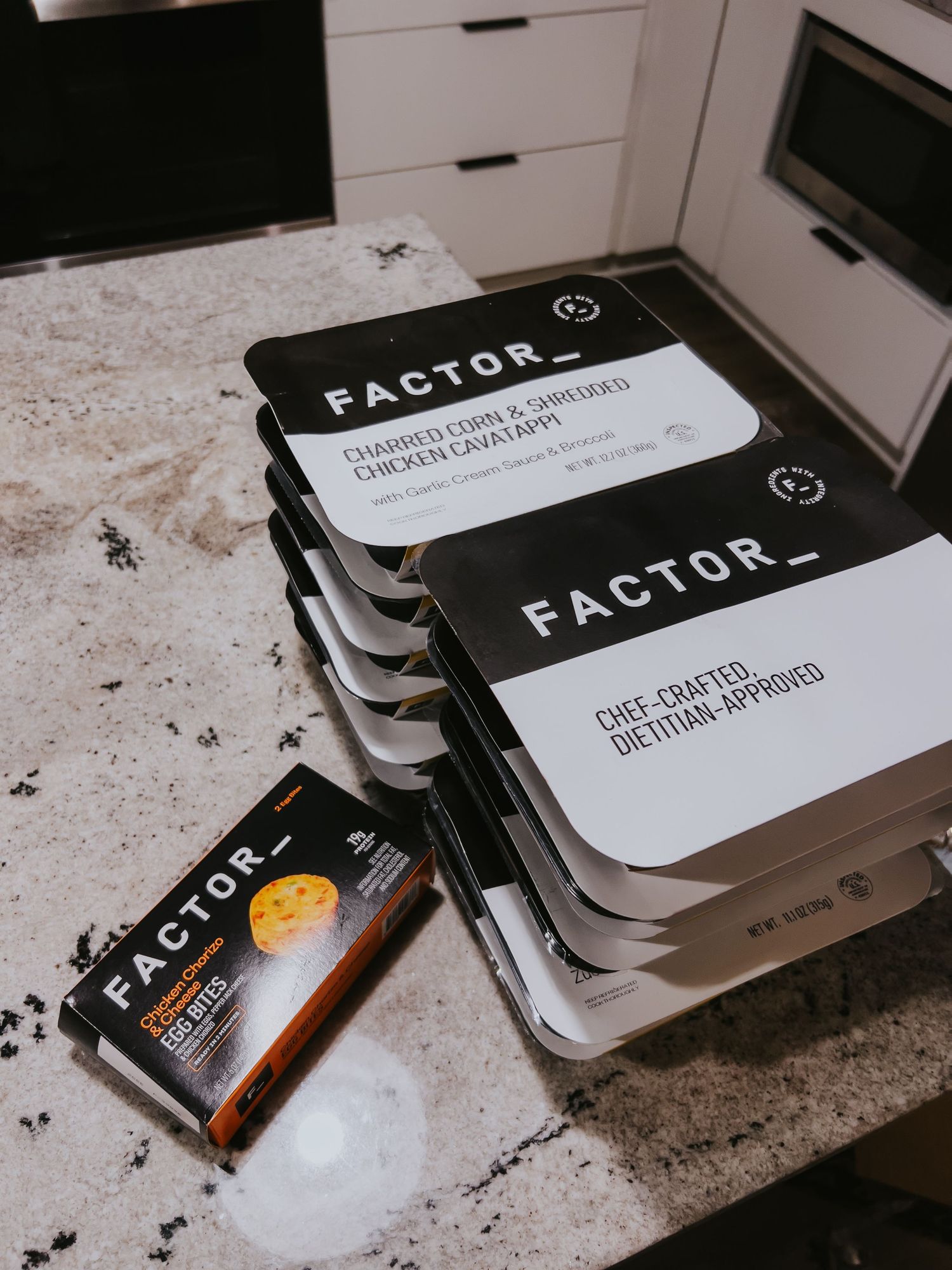 Stacks of black FACTOR meal delivery service boxes and packaging displayed on a white marble countertop.