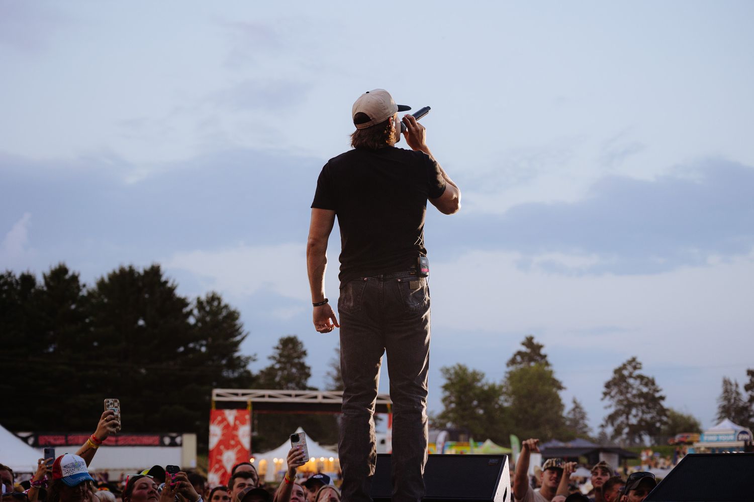 A silhouetted performer stands on stage during an outdoor concert with tents and trees visible in the background.
