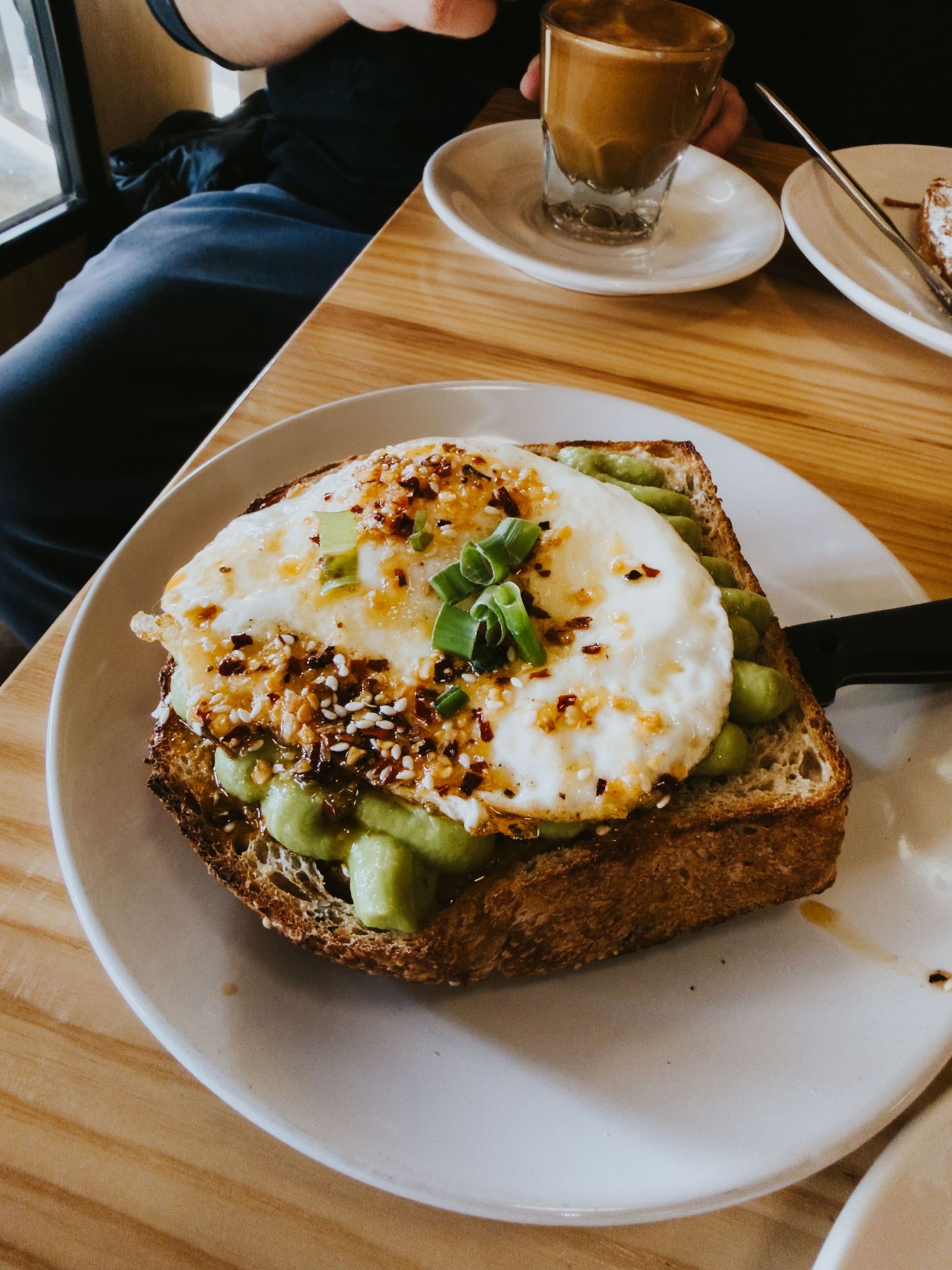 Slices of rustic bread topped with avocado, fried eggs, and sesame seeds served on white plates at a wooden table.