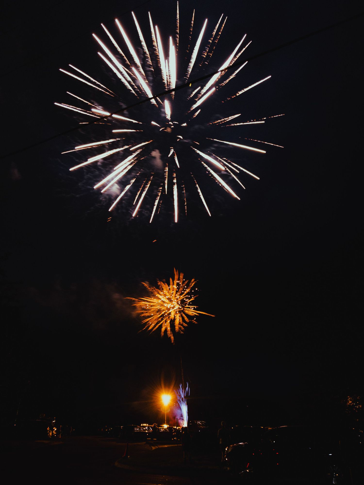 Multiple fireworks bursts illuminate the night sky over a dark cityscape with white and orange pyrotechnic displays.