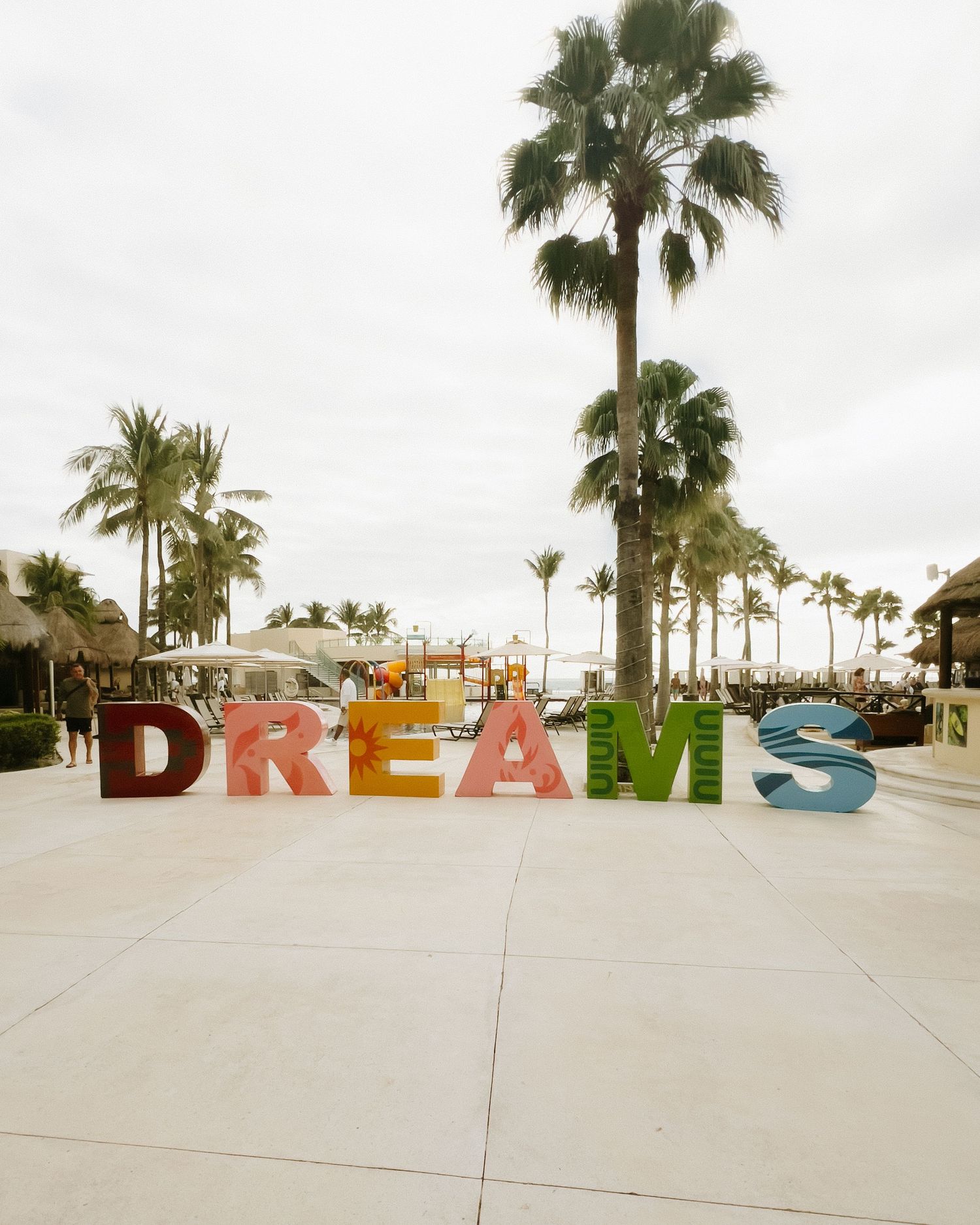 Large colorful DREAMS sign installation displayed on a beachfront promenade with palm trees and cloudy sky.