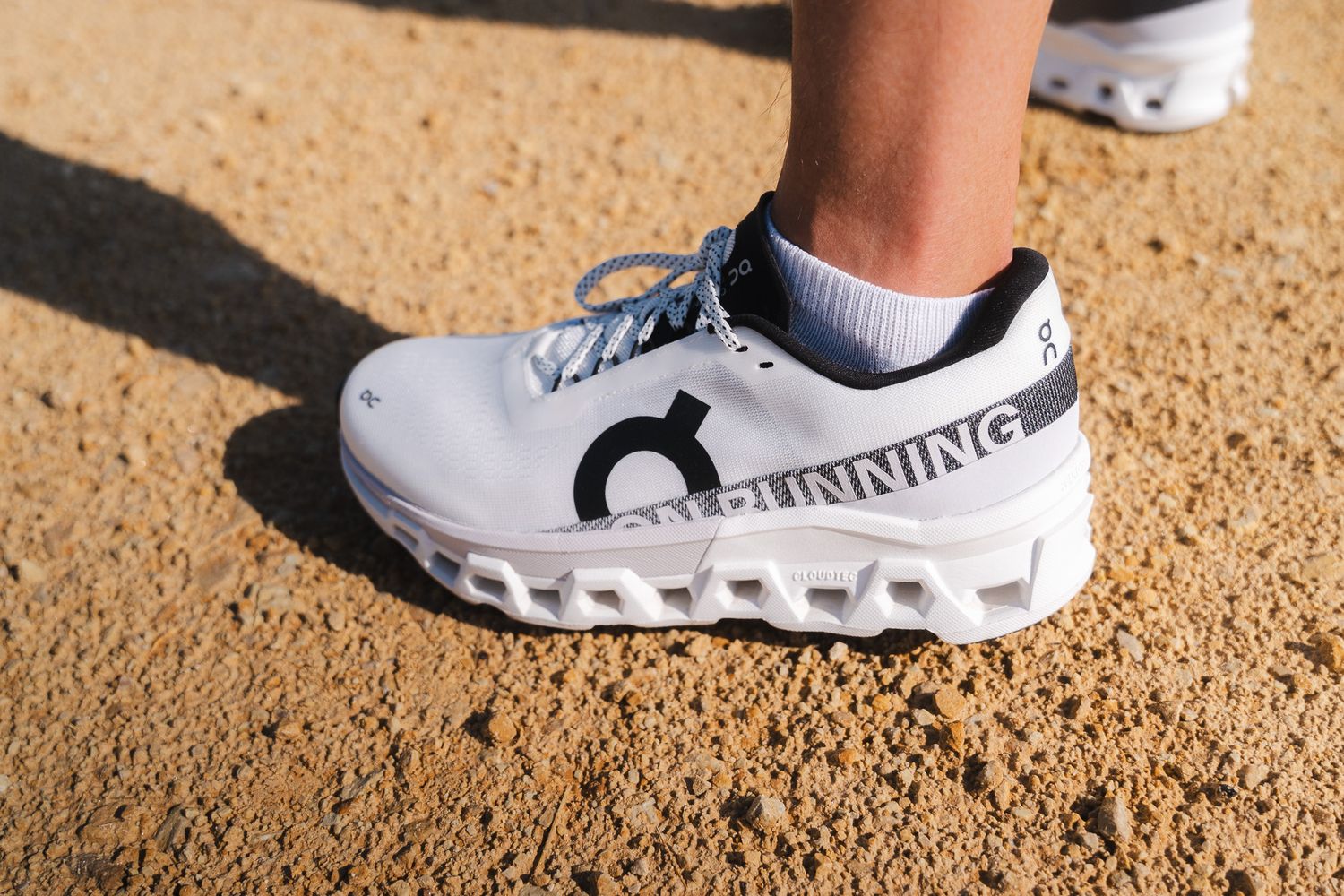 Close-up view of white athletic shoes walking on sandy ground in bright sunlight.