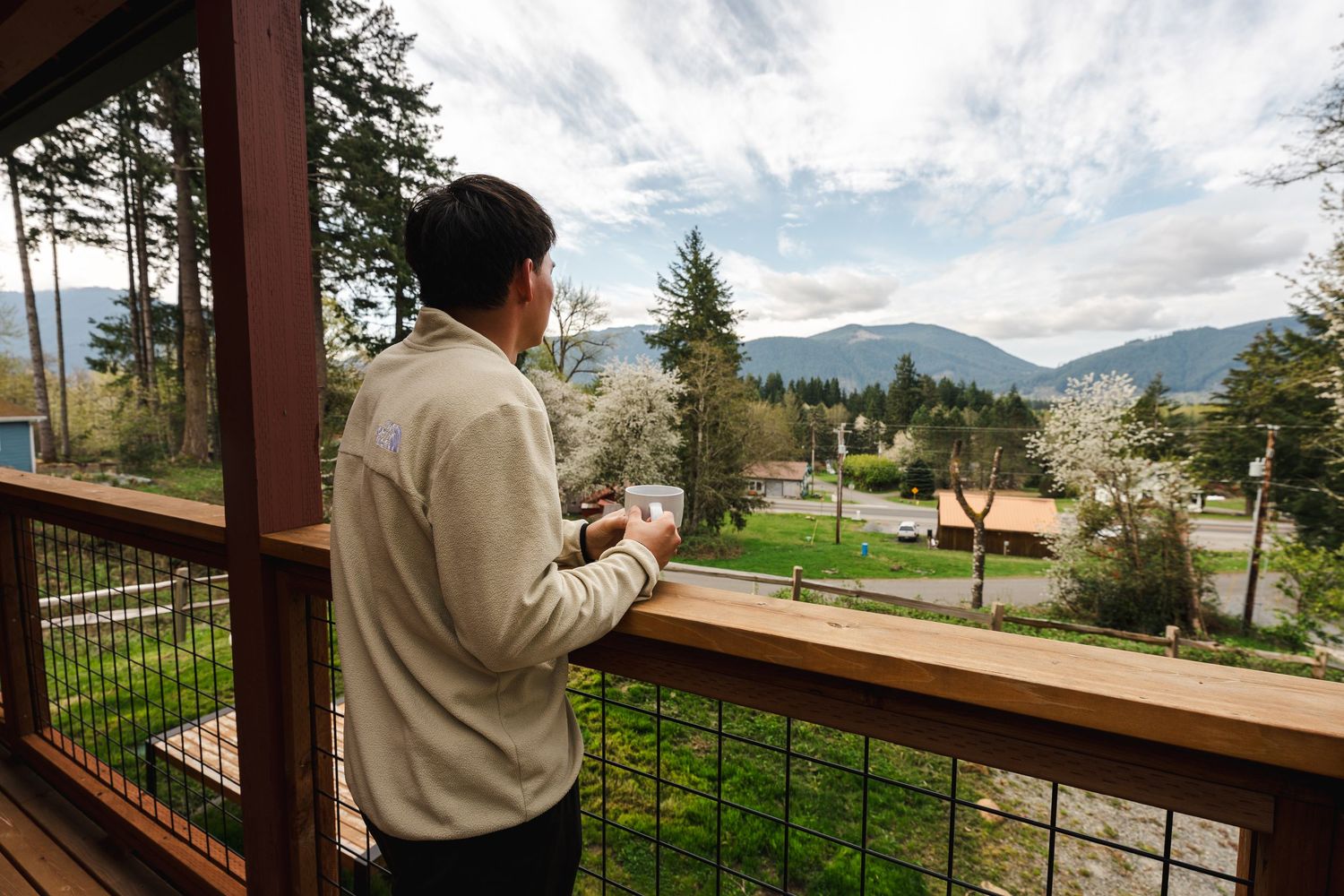 Man standing on a wooden balcony with cable railing, enjoying mountain and valley views while holding a coffee cup.