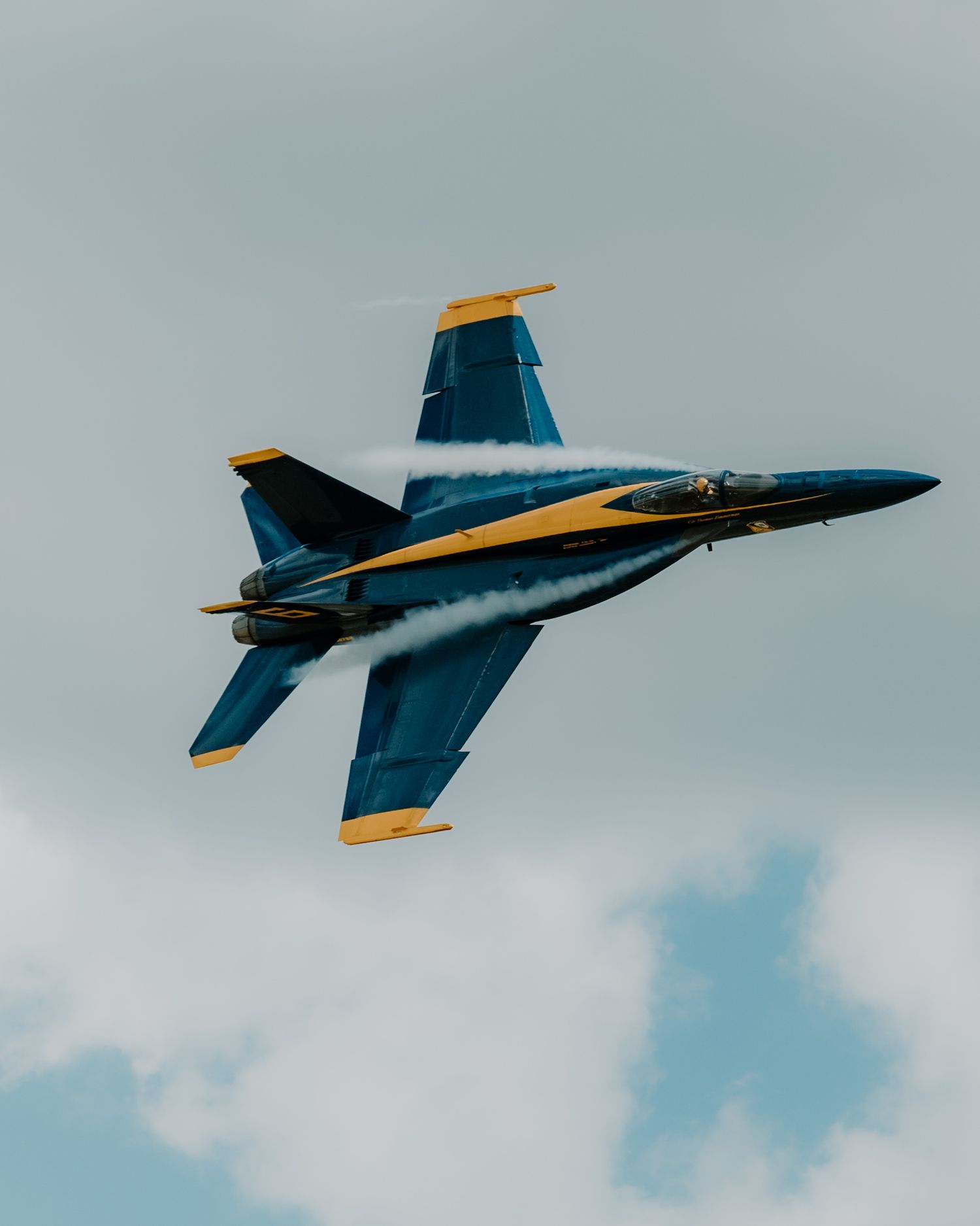 A US Navy Blue Angels F/A-18 Hornet fighter jet performs aerial maneuvers against a cloudy sky with blue and gold livery.