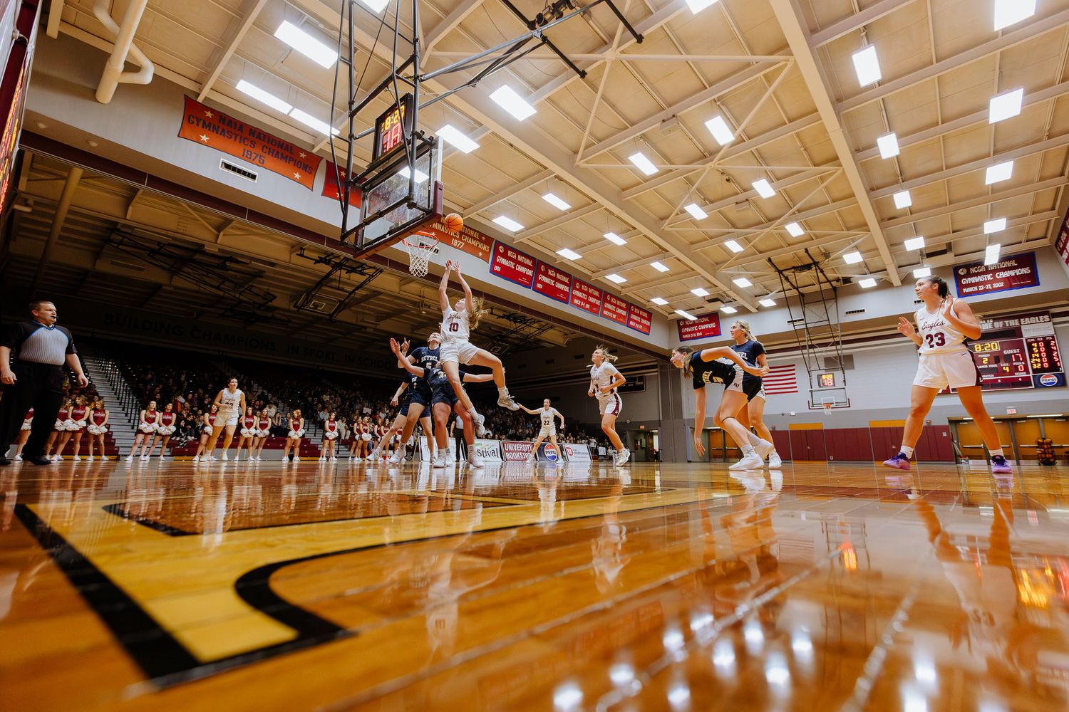 High school basketball players compete on a polished wooden court during an intense game in a brightly lit gymnasium.