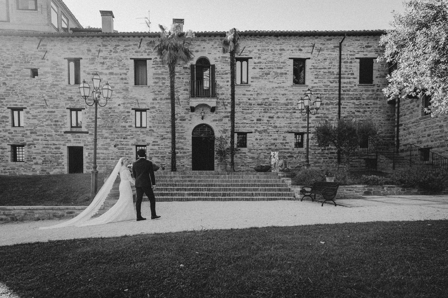 Brautpaar geht Hand in Hand auf die Villa Teloni zu, Rückenansicht mit langem Schleier