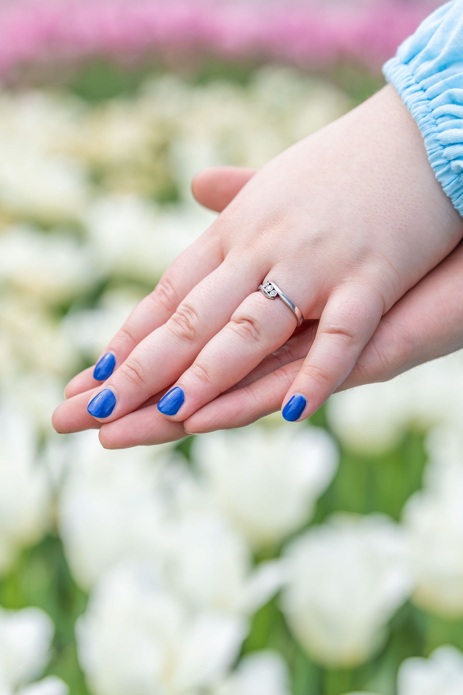 Hand with vibrant blue nail polish and engagement ring shown against a blurry background of white and pink flowers.