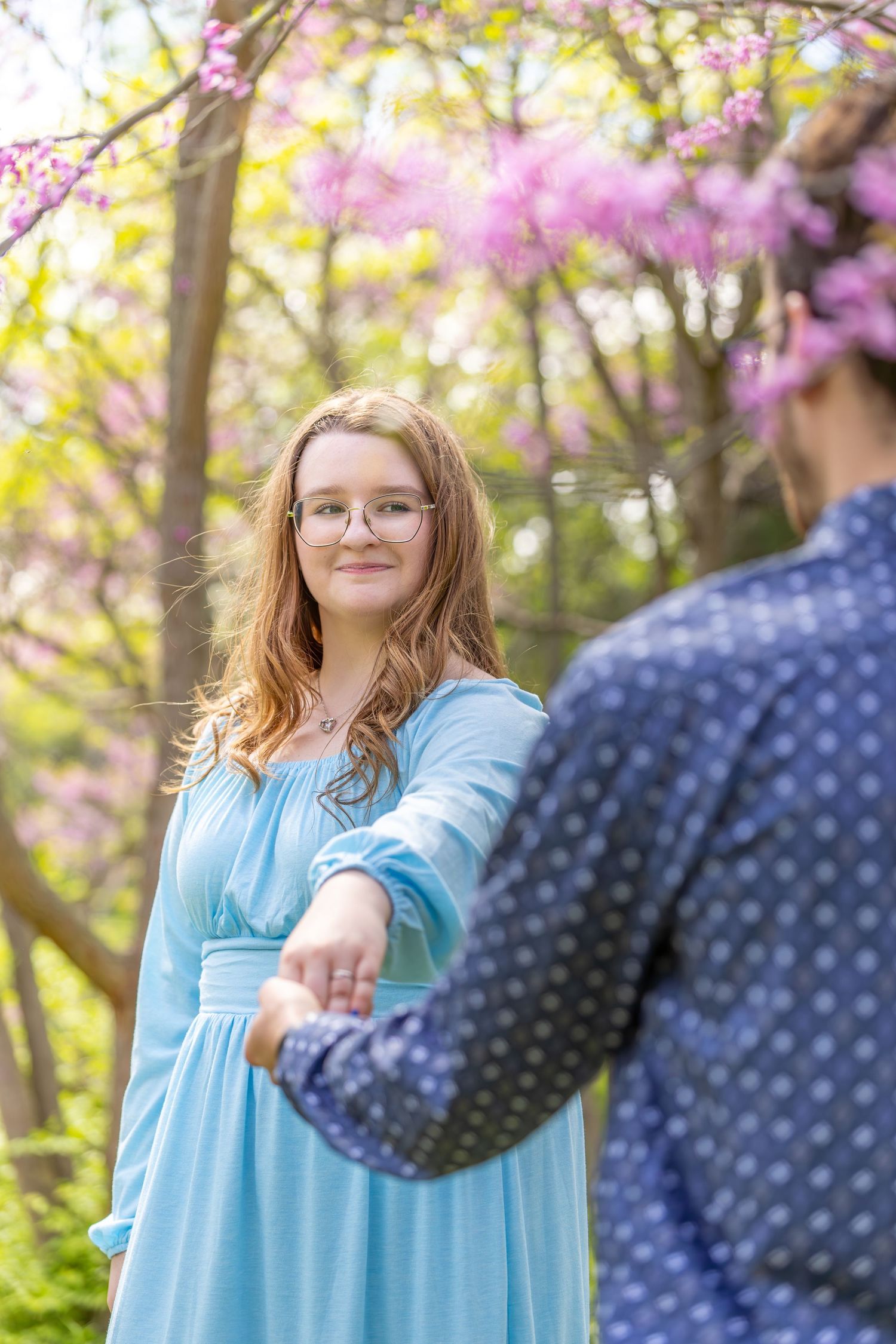 A couple holds hands during a spring photoshoot with blooming cherry blossoms and soft bokeh in the background.