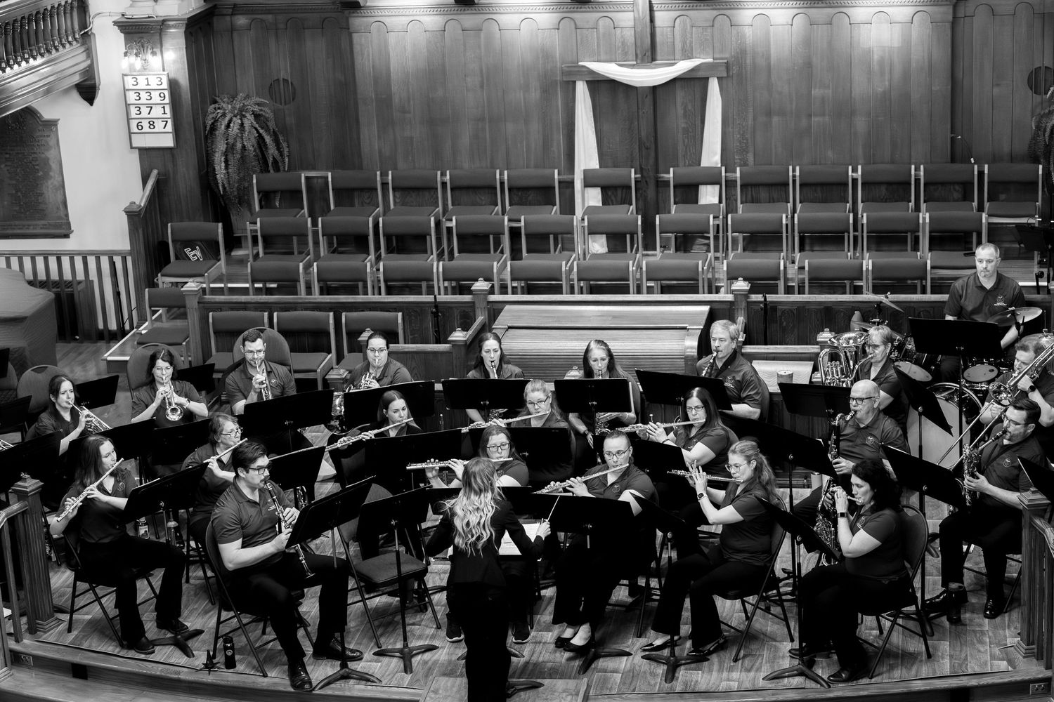 Black and white photo of a community orchestra performing on stage with musicians seated at music stands and empty chairs behind them.