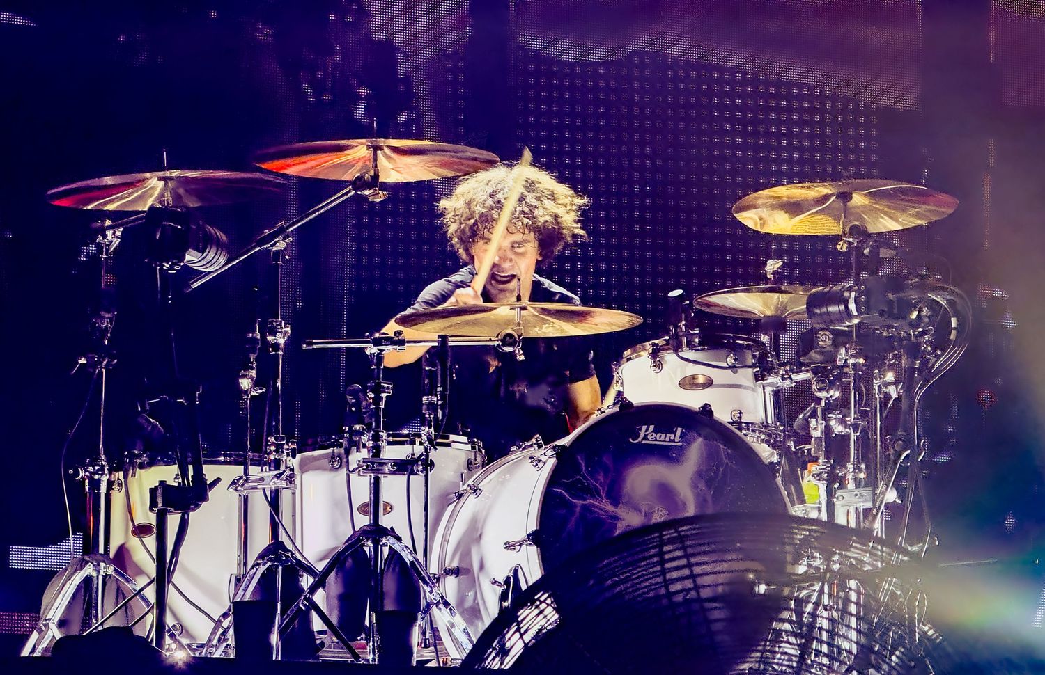A drummer performs on stage with a large drum kit setup illuminated by stage lighting during a live performance.