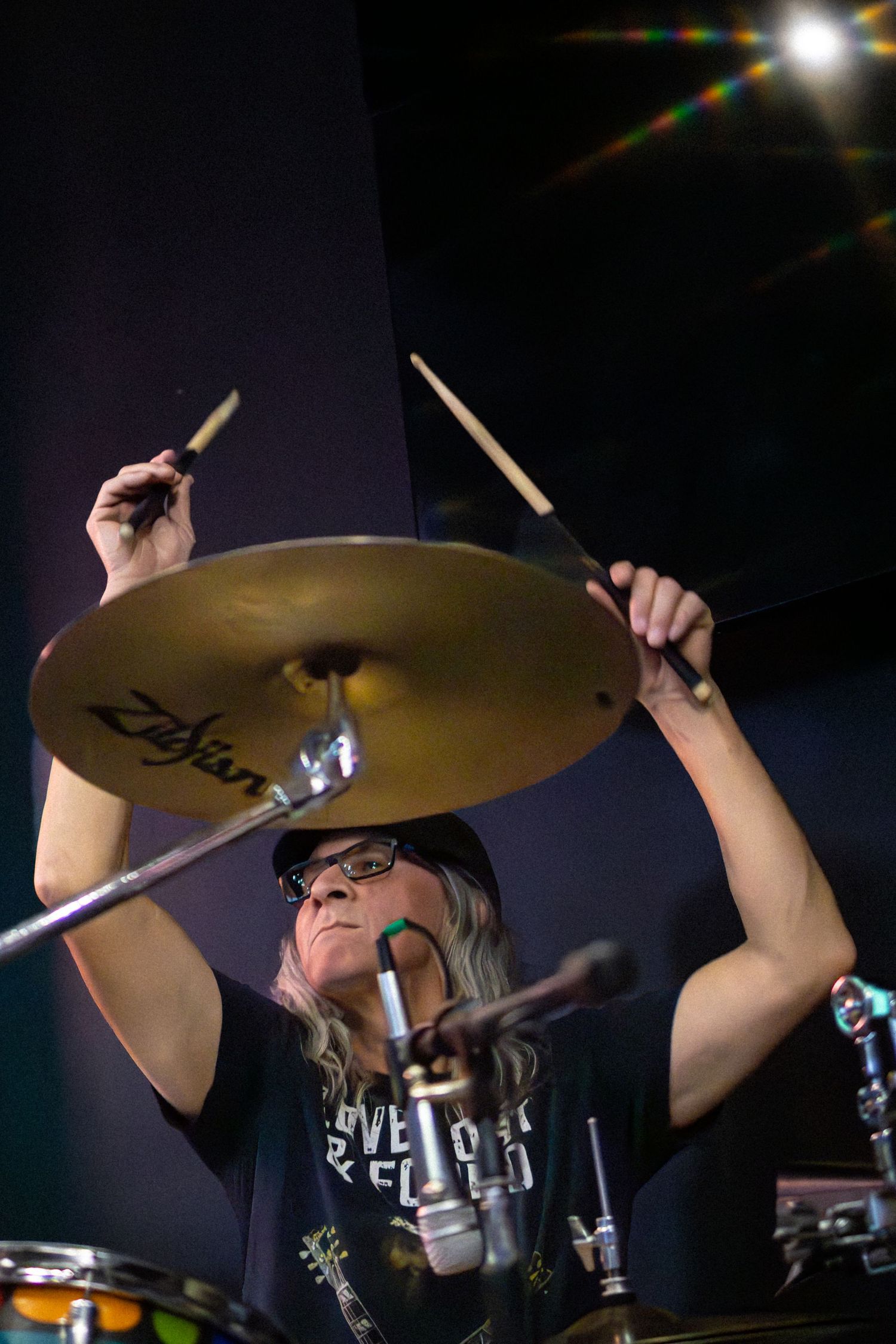 Drummer performing on stage with cymbals, drumsticks raised in motion during a dynamic performance in dark lighting.