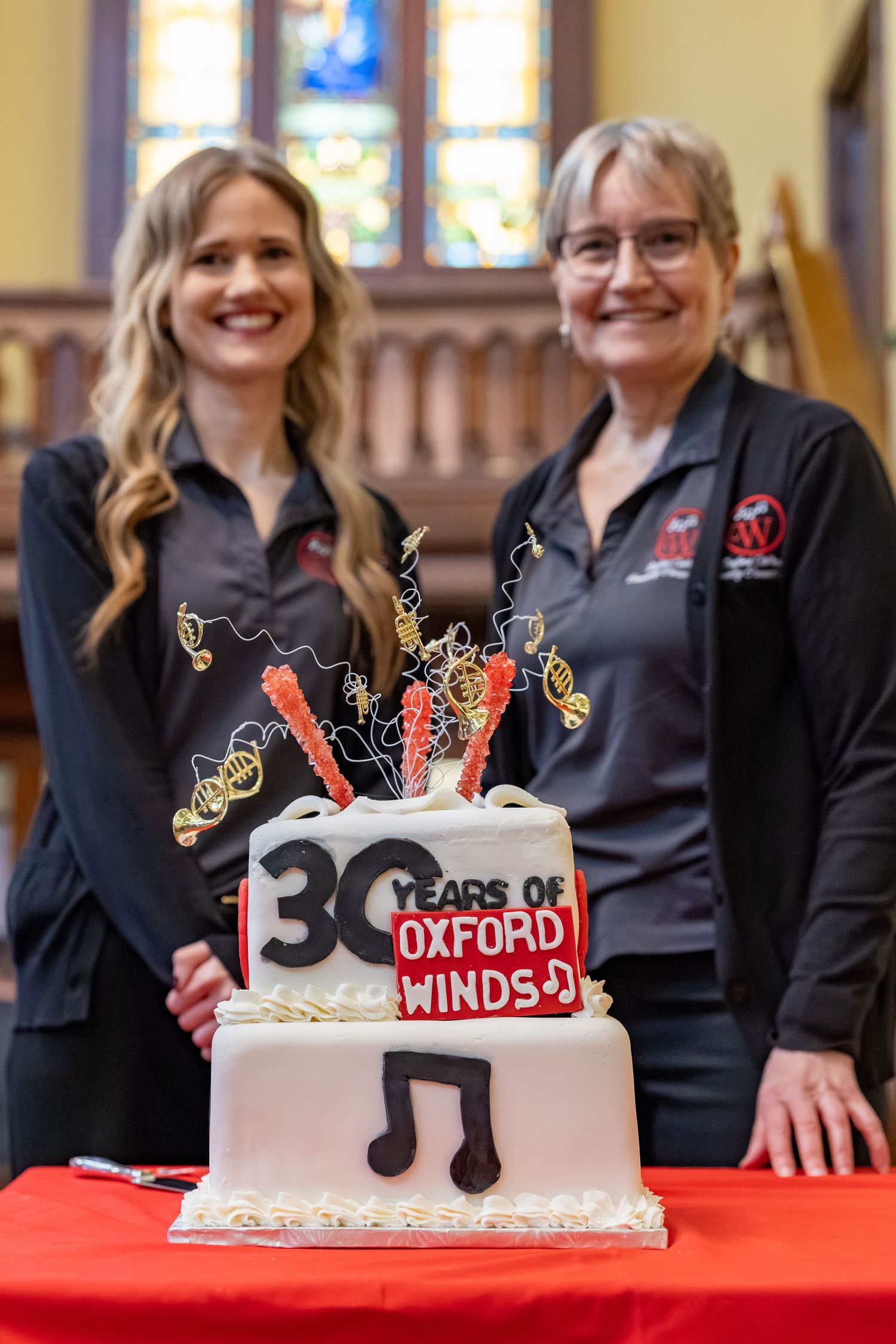 Two smiling women in matching dark polos stand behind a 30 Years of Oxford Winds anniversary cake decorated with music notes.