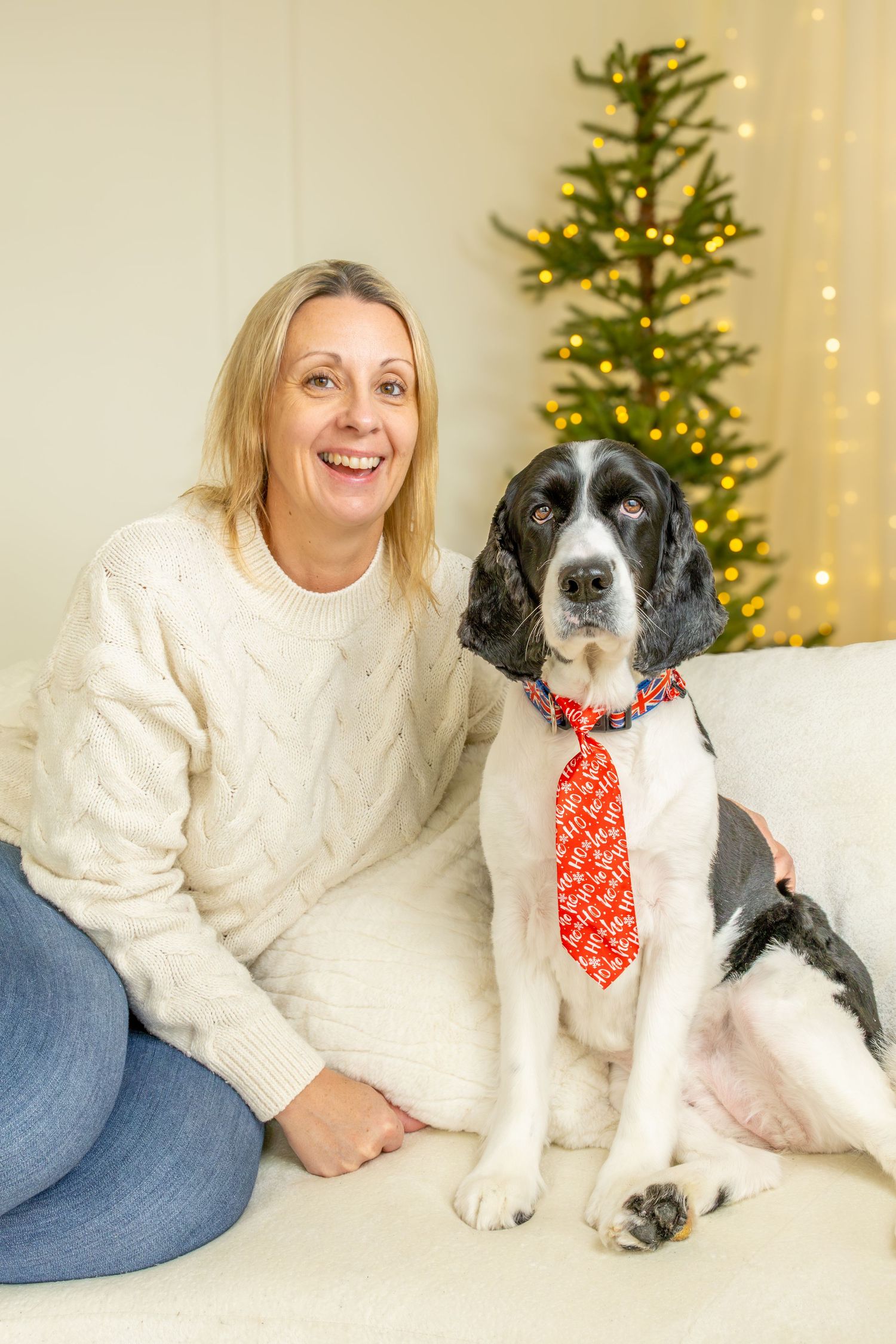 Smiling woman in a cream cable-knit sweater sits on a sofa beside a springer spaniel wearing a red tie, with a lit Christmas tree behind.