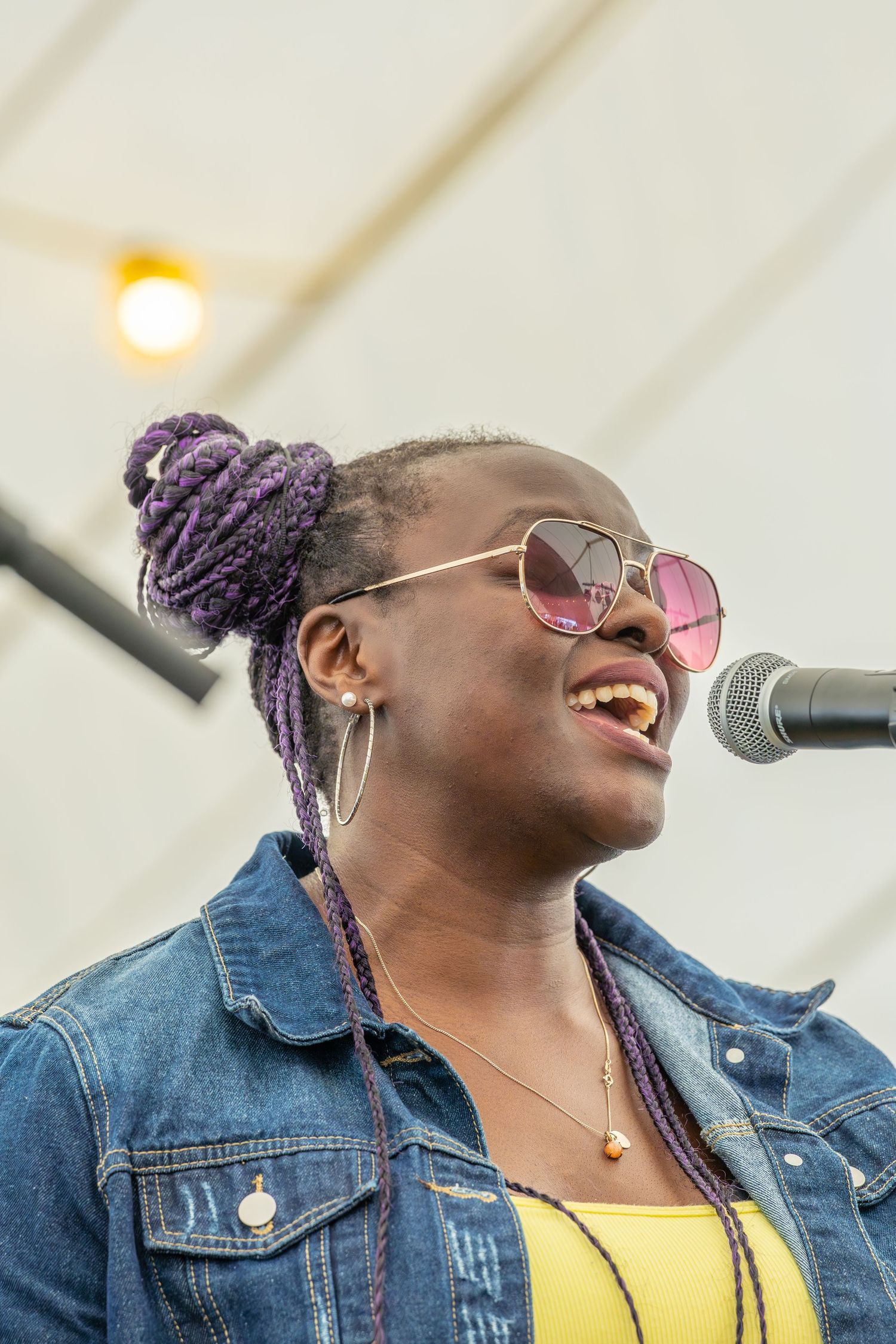 A performer wearing a denim jacket and pink sunglasses smiles joyfully while singing into a microphone at an outdoor event.